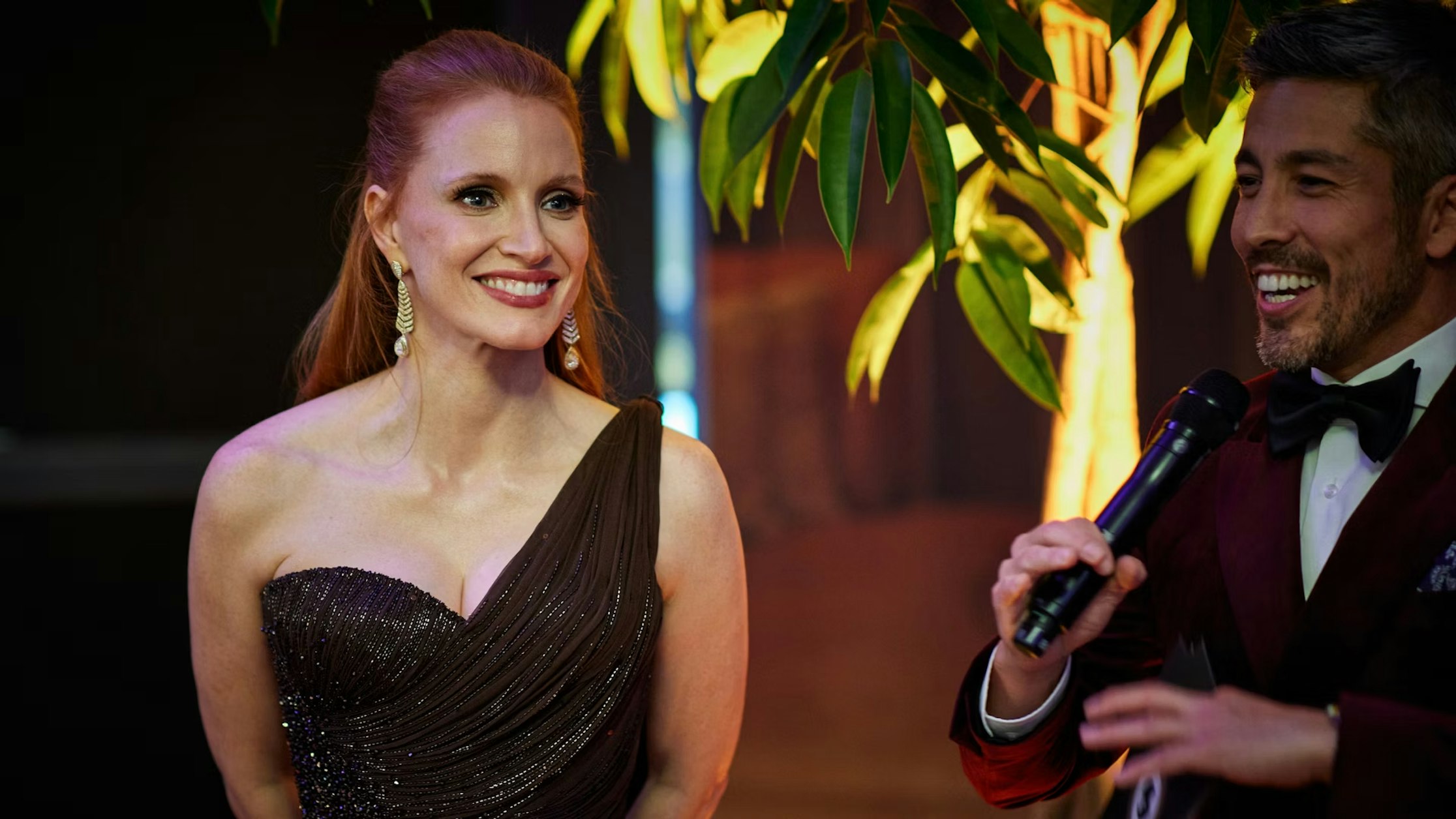 Eine Frau in einem glitzernden, schulterfreien schwarzen Kleid steht lächelnd neben einem Mann im Smoking, der ein Mikrofon hält. Sie scheinen sich auf einer formellen Veranstaltung zu befinden, mit viel Grün und sanfter Beleuchtung im Hintergrund.
