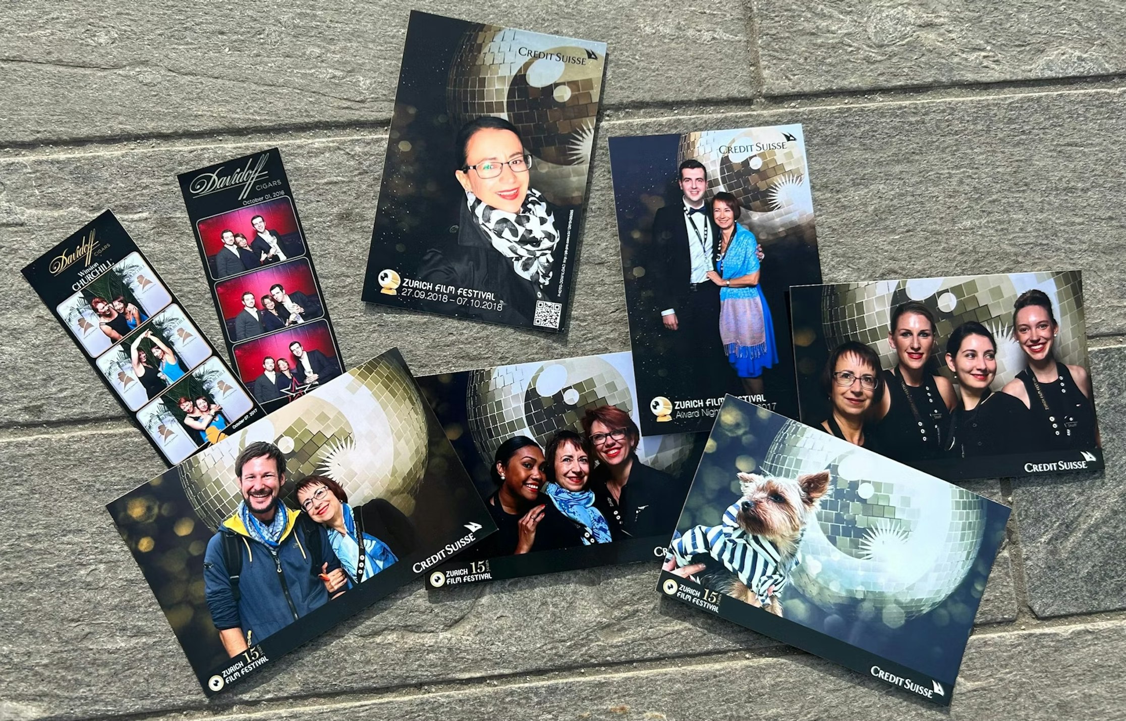 Eine Sammlung ausgedruckter Fotoautomatenbilder von einer Veranstaltung ist auf einer Steinfläche angeordnet. Zu sehen sind Personen, die paarweise oder in Gruppen posieren, einige in Kostümen, und auf einem Foto ist ein Hund zu sehen. Auf den meisten Fotos ist eine Discokugel im Hintergrund zu sehen.