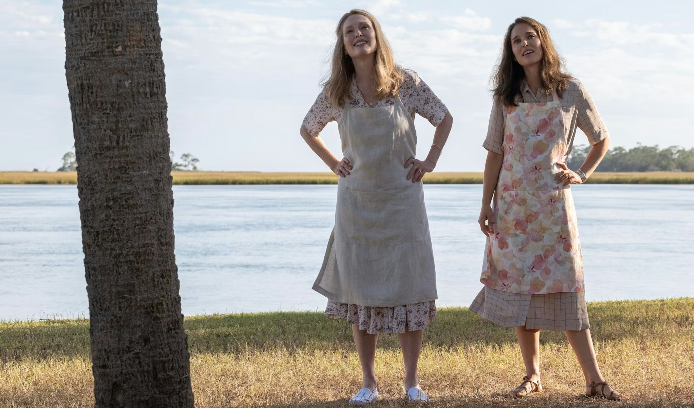 Zwei Frauen mit Schürzen stehen an einem sonnigen Tag im Gras neben einem Baum, im Hintergrund ein Fluss und Felder. Beide lächeln und wirken entspannt und genießen die Natur.