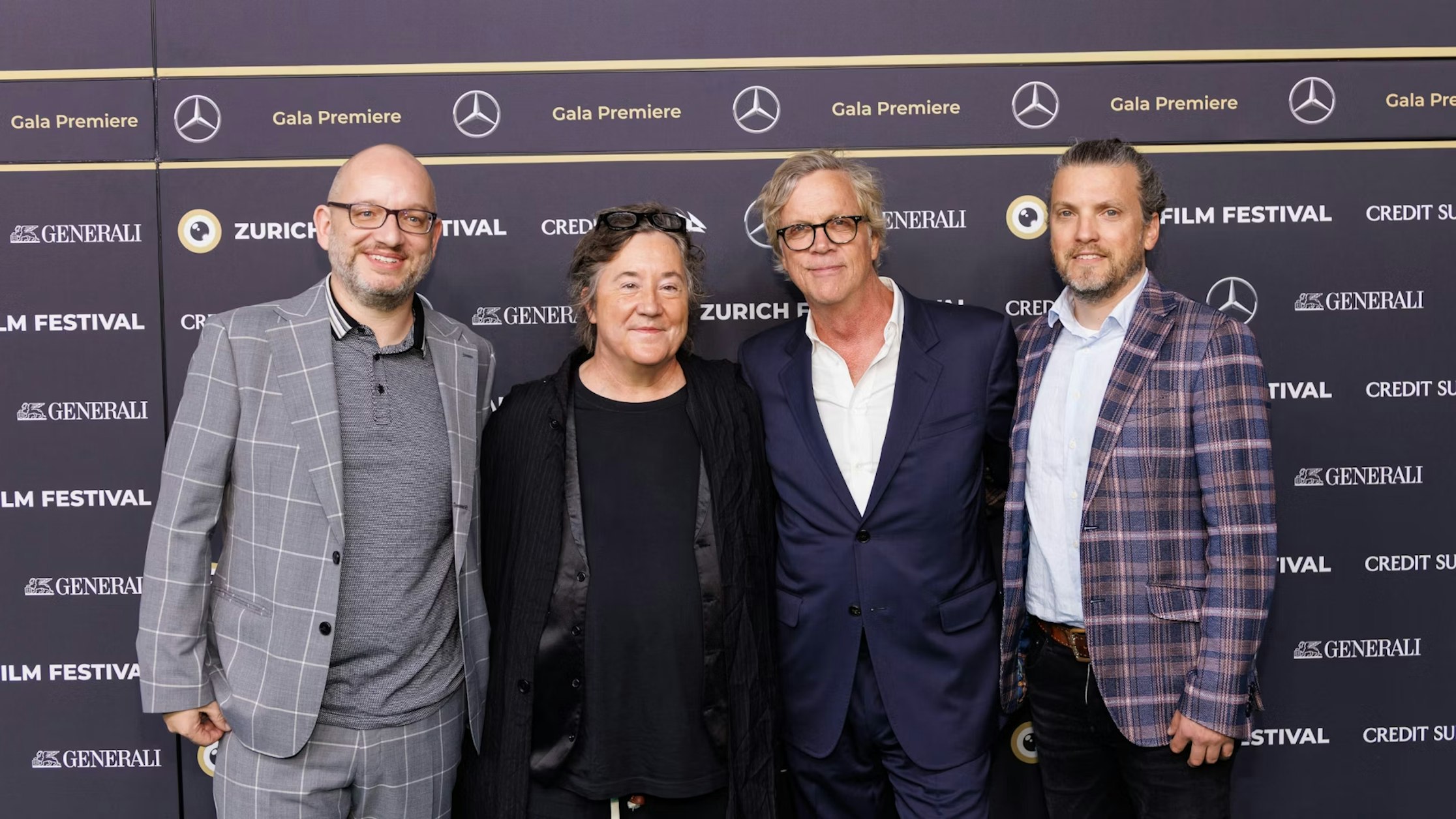 Vier Männer in Anzug und Jackett stehen lächelnd vor einer Bühnenkulisse des Zurich Film Festival und posieren für ein Gruppenfoto bei einer Gala-Premierenveranstaltung.