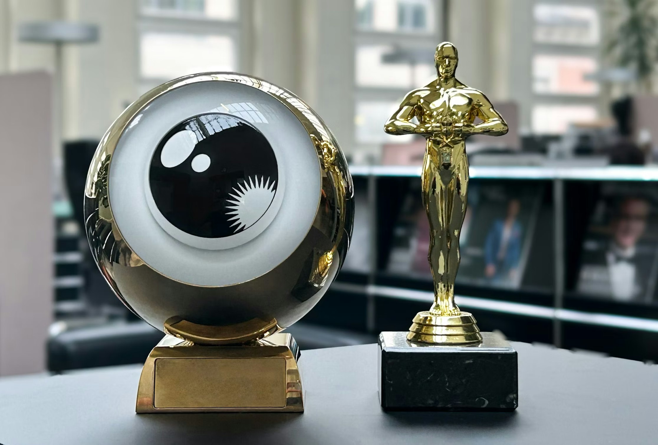 Eine goldene Trophäe in Form eines Auges neben einer goldenen Oscar-ähnlichen Statuette auf einem schwarzen Marmorsockel, ausgestellt auf einem Tisch in einem modernen, gut beleuchteten Büroumfeld.