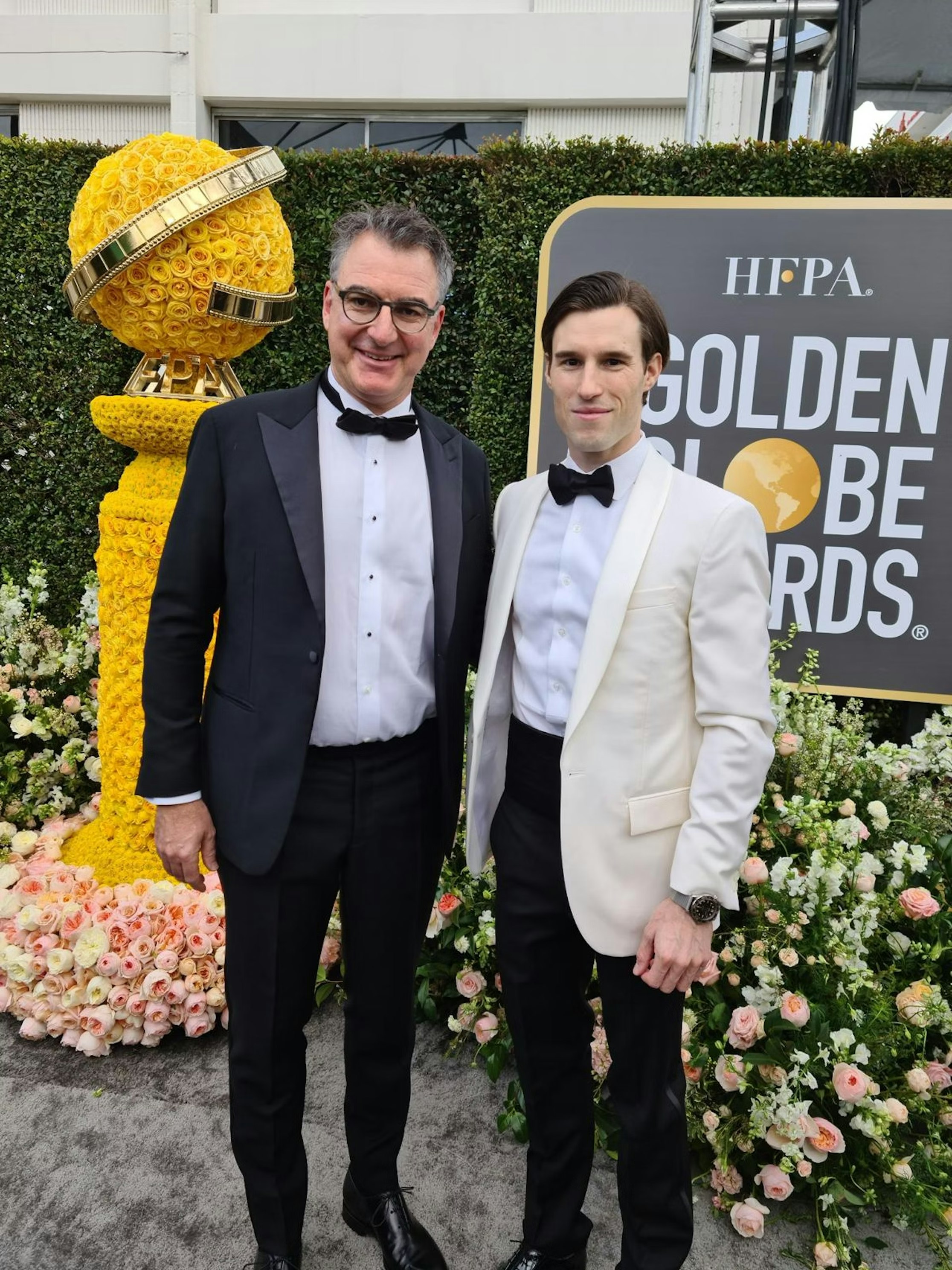 Zwei Männer im Smoking stehen auf einem roten Teppich vor einem Golden Globe Awards-Schild und einer großen Golden Globe-Statue aus gelben Blumen, umgeben von Blumenarrangements. Beide lächeln und posieren für das Foto.