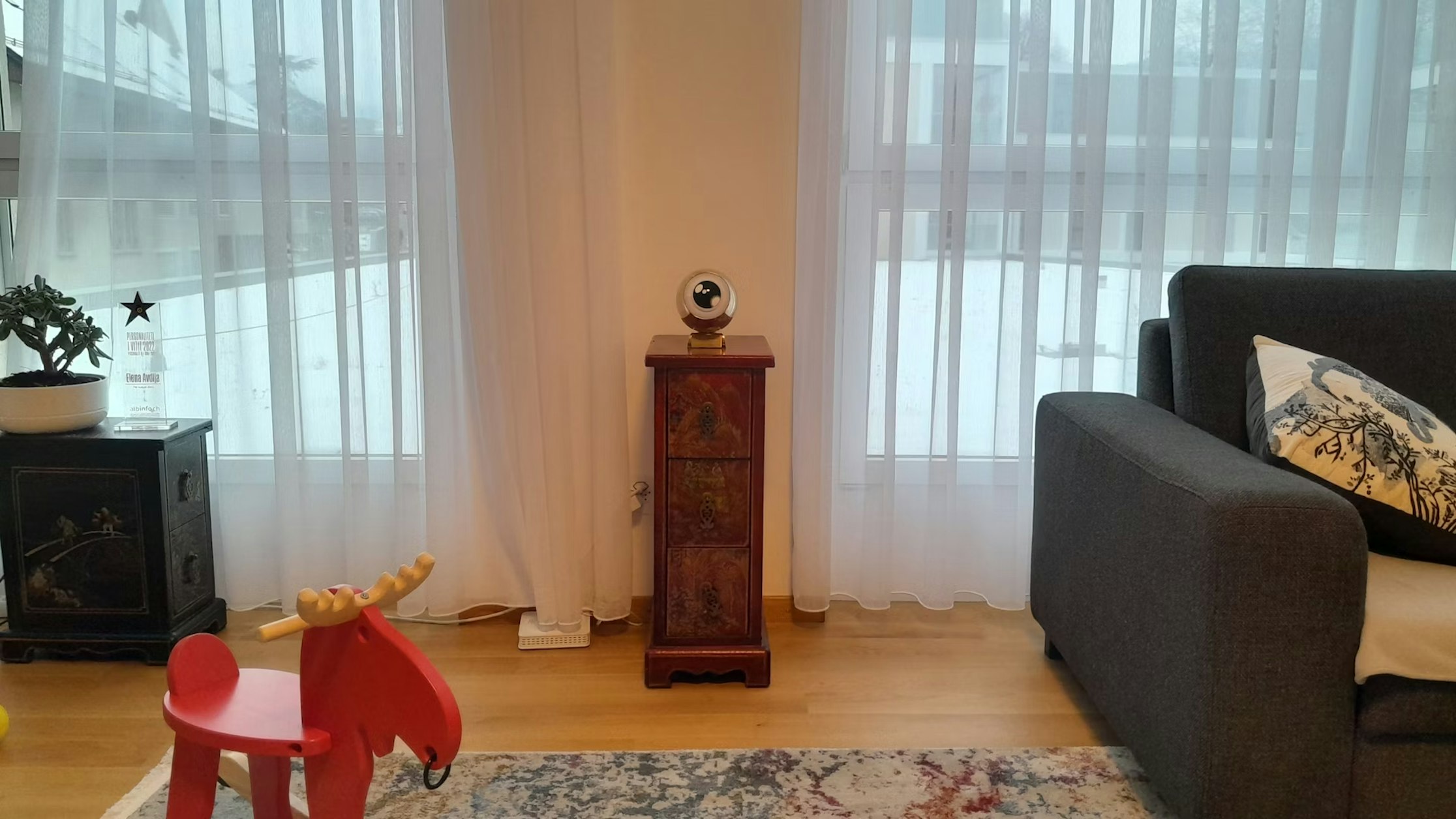 Ein gemütliches Wohnzimmer mit einem grauen Sofa, einem roten Spielzeug-Elch aus Holz, einem kleinen Schrank mit einem Globus darauf, durchsichtigen weißen Vorhängen und vor den großen Fenstern ist Schnee zu sehen.