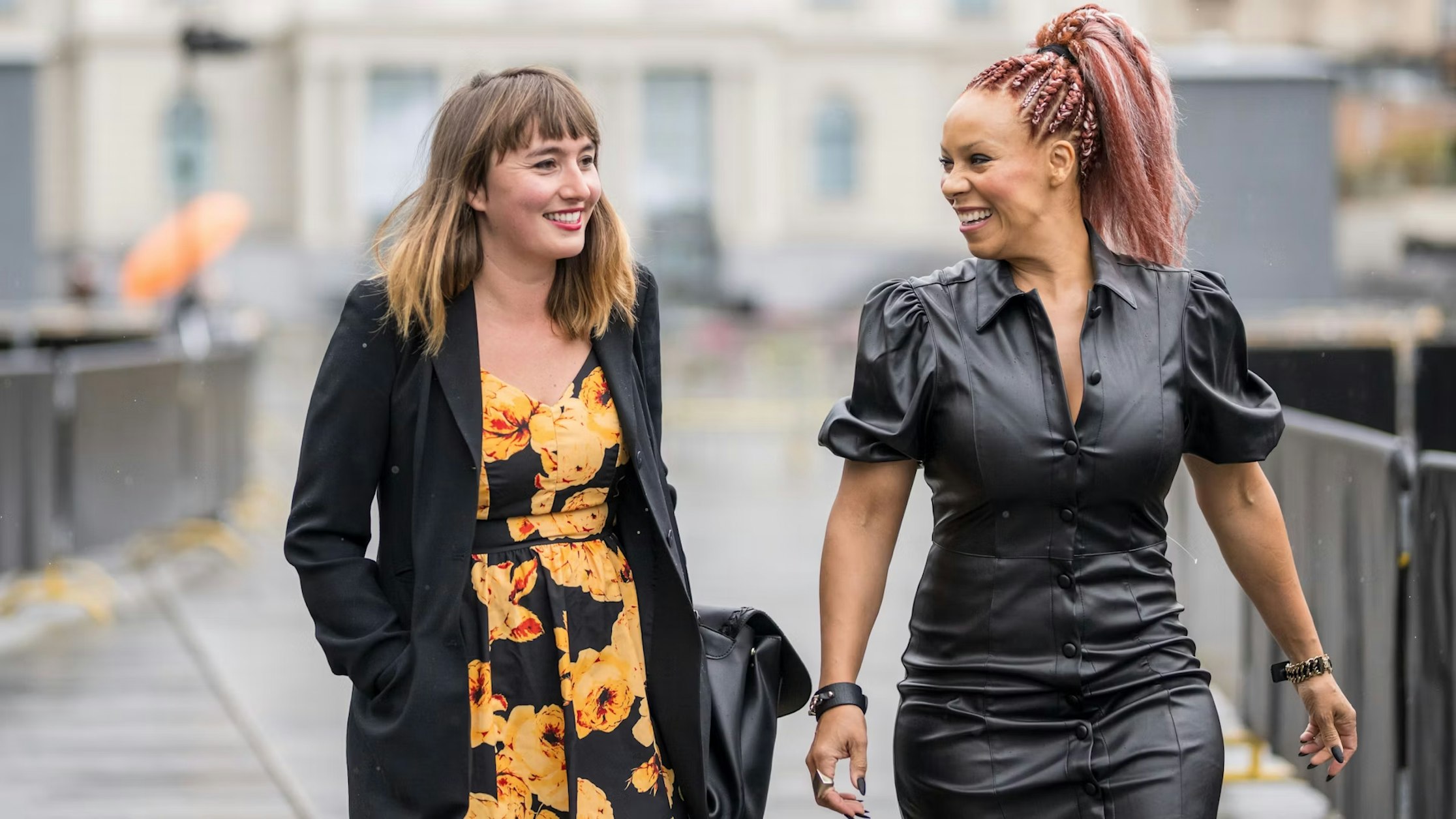 Zwei Frauen gehen an einem regnerischen Tag gemeinsam im Freien spazieren, lächeln und unterhalten sich. Die eine trägt ein schwarzes Blumenkleid und einen Blazer, die andere ein schwarzes Lederkleid mit Puffärmeln und geflochtenen Haaren. Der Hintergrund ist leicht unscharf.