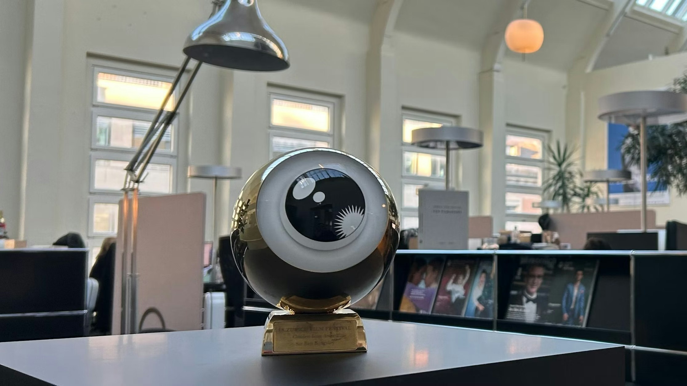 Eine goldene Trophäe in Form eines übergroßen Augapfels steht auf einem Schreibtisch in einem modernen Büroraum. Im Hintergrund sind große Fenster, Lampen und Pflanzen zu sehen.