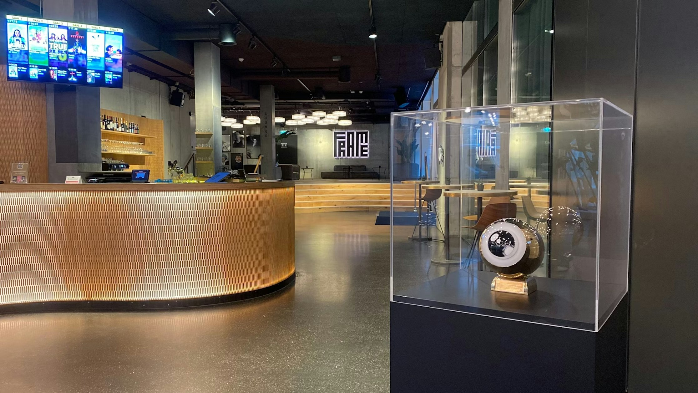 Modernes Kinofoyer mit einem geschwungenen, gefliesten Tresen links, digitalen Kinoleinwänden darüber und einer Trophäenvitrine rechts, in der eine Auszeichnung ausgestellt ist. Im Hintergrund ist an der Rückwand „FAME“ zu sehen.