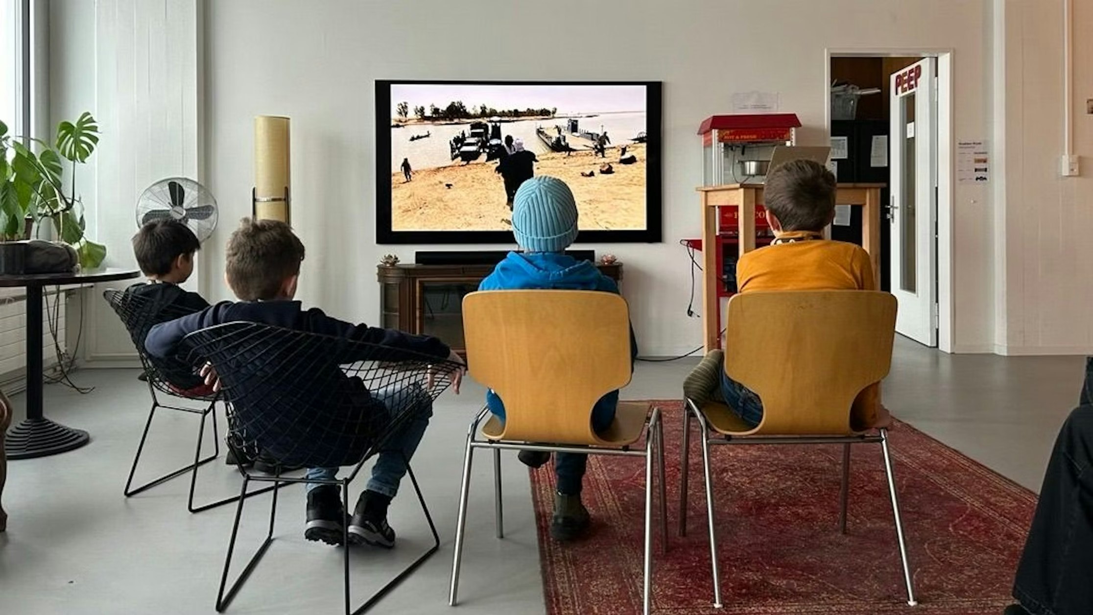 Vier Kinder sitzen auf Stühlen vor einem Fernseher und schauen sich eine Szene mit Fahrzeugen in einer Sandlandschaft an. Der Raum ist mit einem roten Teppich, Pflanzen und einer Popcornmaschine ausgestattet. Die Atmosphäre erinnert an ein entspanntes gemeinsames Filmschauen.