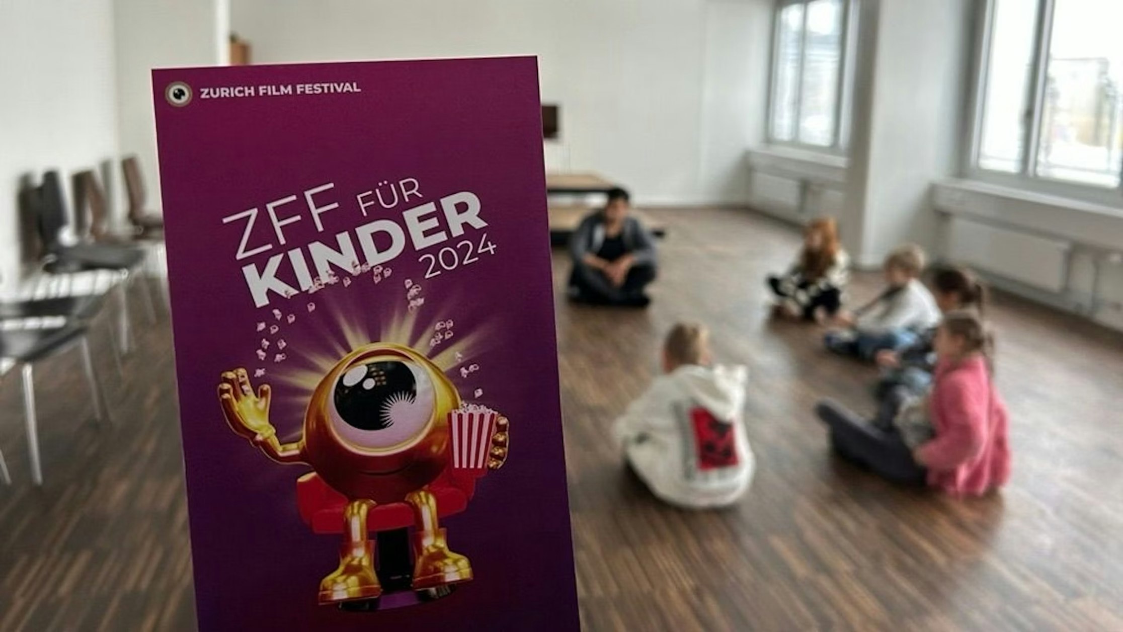 Eine Hand hält einen lila Flyer für ZFF für Kinder 2024 mit einem Cartoon-Maskottchen. Im Hintergrund sitzen Kinder im Kreis auf dem Boden eines grosszügigen, hellen Raumes, an der Wand sind Stühle aufgereiht.