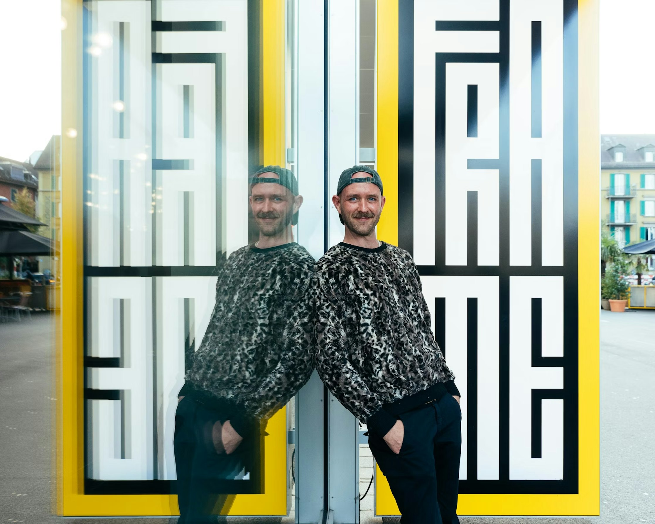 Ein Mann in gemustertem Pullover und Mütze lehnt an einer Glaswand, die eine Spiegelung erzeugt. Hinter ihm ist ein auffälliges schwarz-weißes geometrisches Muster mit gelbem Rahmen und dem Wort FRAME zu sehen. Im Hintergrund ist eine städtische Außenkulisse zu sehen.