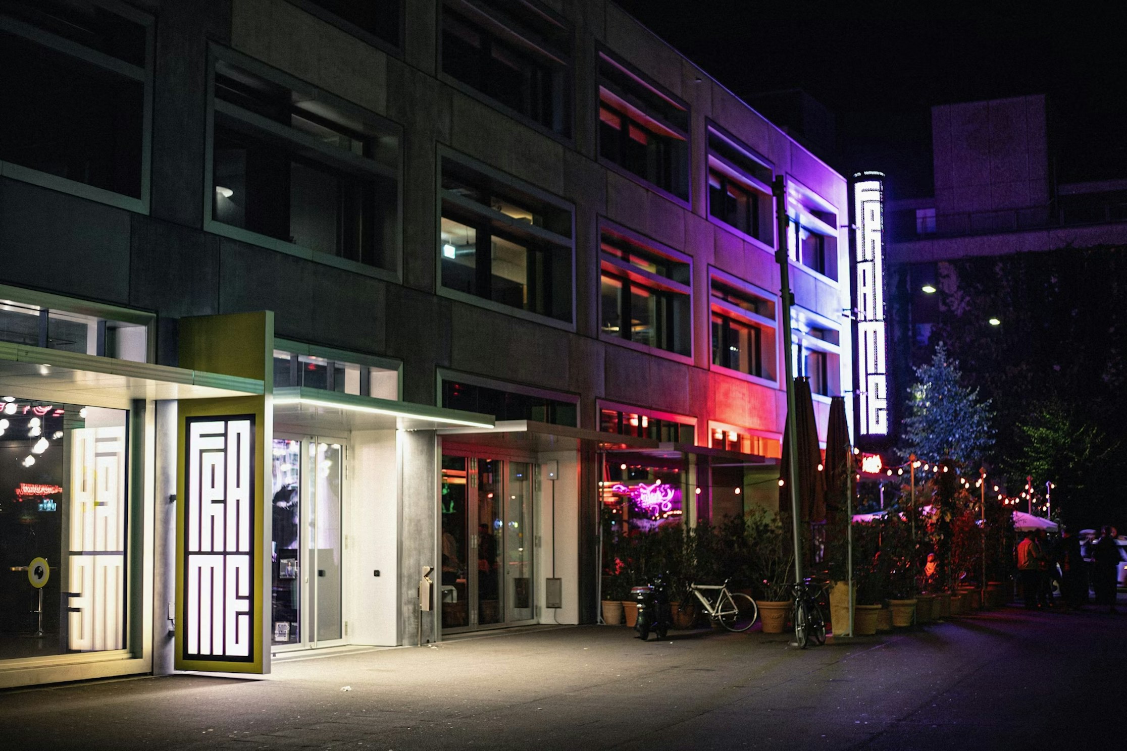 Ein modernes Gebäude bei Nacht mit großen Fenstern und leuchtenden „FRAME“-Neonschildern. Helle, bunte Lichter erhellen den Sitzbereich im Freien, wo Fahrräder abgestellt sind und sich Menschen unter Sonnenschirmen versammeln.