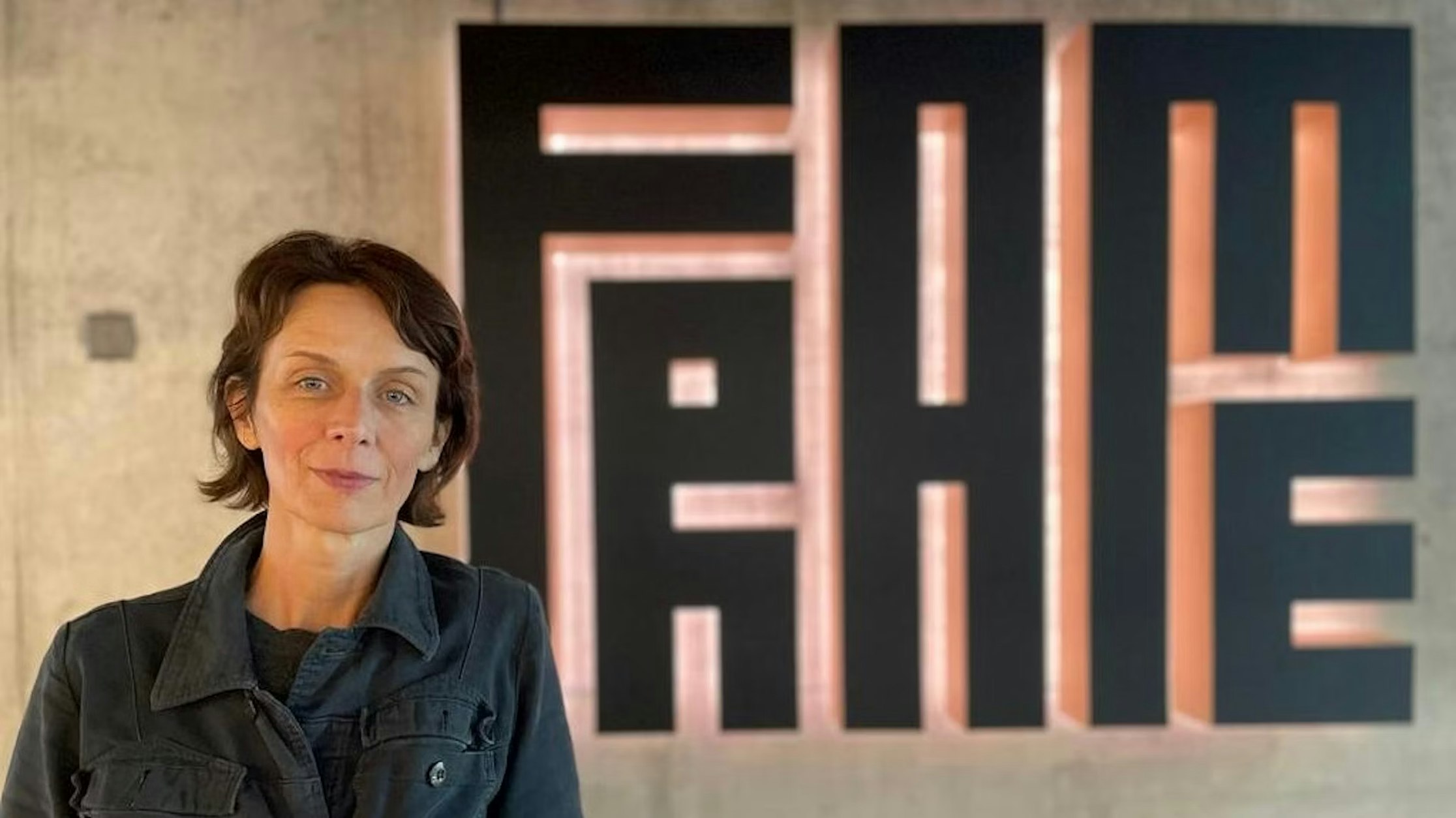 Eine Frau mit kurzen braunen Haaren und einer dunklen Jacke steht vor einem großen Wandschild, auf dem in fetten, geometrischen schwarzen Buchstaben „FAME“ steht.