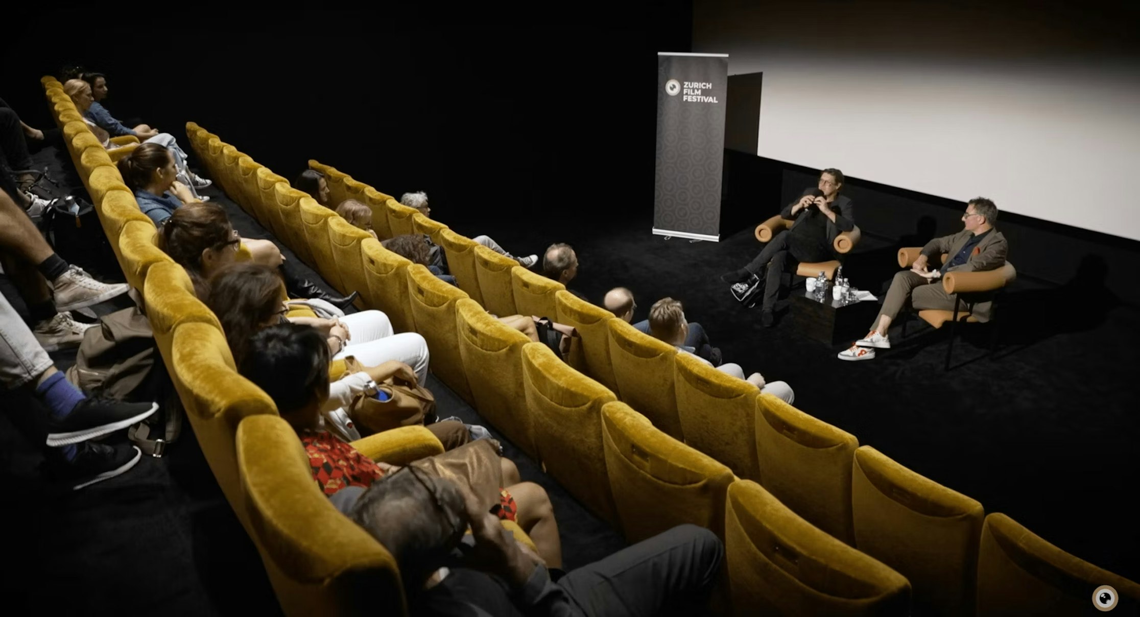 Ein kleines Publikum sitzt auf senfgelben Stühlen gegenüber zwei Personen, die bei einer Filmfestivalveranstaltung auf der Bühne vor einer Leinwand eine Diskussion führen.