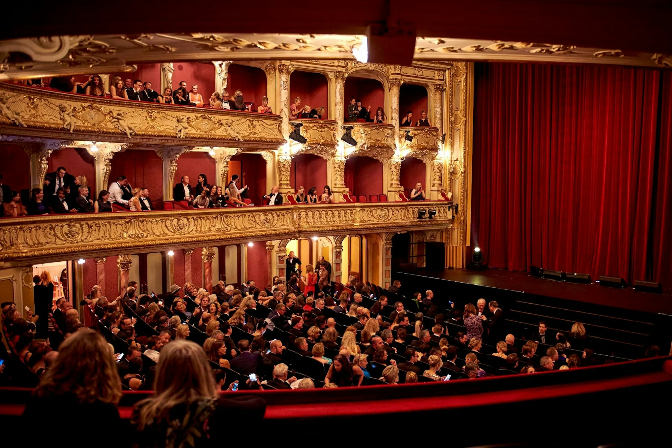 Ein großes Publikum sitzt in einem prächtigen, kunstvoll verzierten Theater mit Balkonen und roten Samtvorhängen und wartet auf den Beginn der Vorstellung. Das Theater ist mit goldenen Verzierungen verziert und hat mehrere Ebenen voller Menschen.
