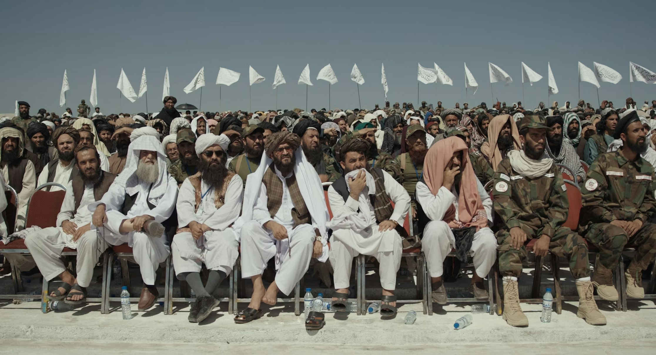 Eine große Gruppe von Männern, einige in traditioneller Kleidung und andere in Tarnuniformen, sitzt in Reihen im Freien, hinter ihnen wehen unter einem klaren Himmel weiße Fahnen.