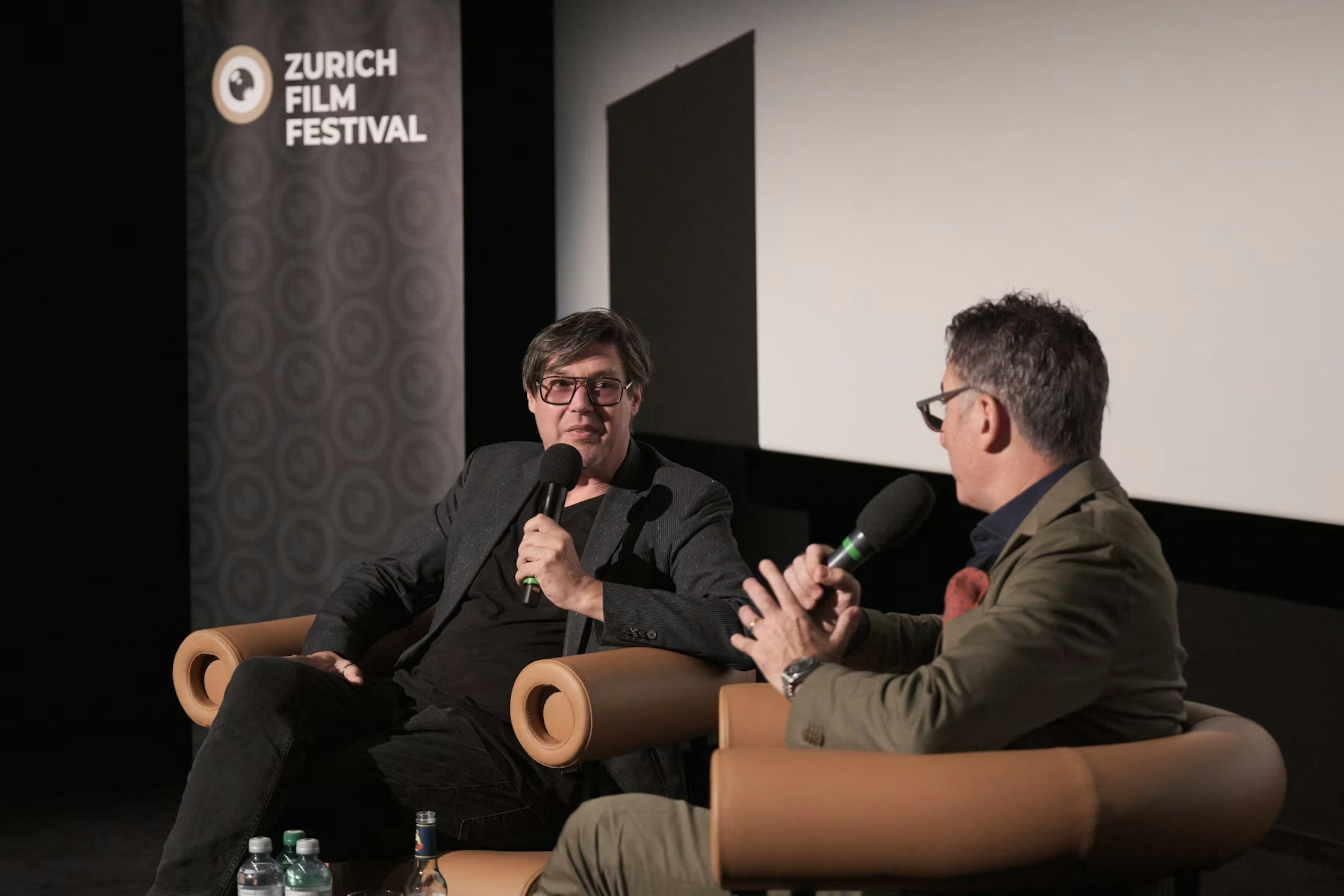 Beim Zurich Film Festival sitzen zwei Männer in Sesseln auf der Bühne und sprechen in Mikrofone. Im Hintergrund sind ein Festivalbanner und eine Projektionsleinwand zu sehen.