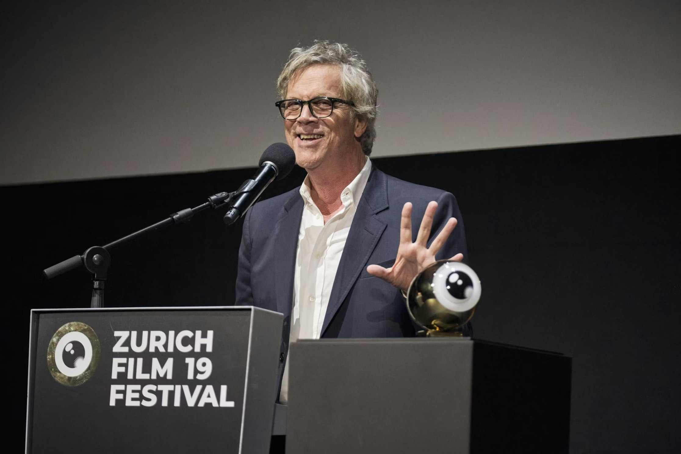 Ein Mann in Anzug und Brille spricht in ein Mikrofon auf einem Podium mit der Aufschrift „Zürich Film 19 Festival“. Er lächelt und gestikuliert mit einer Hand. Auf dem Podium steht eine Trophäe in Form eines Auges.