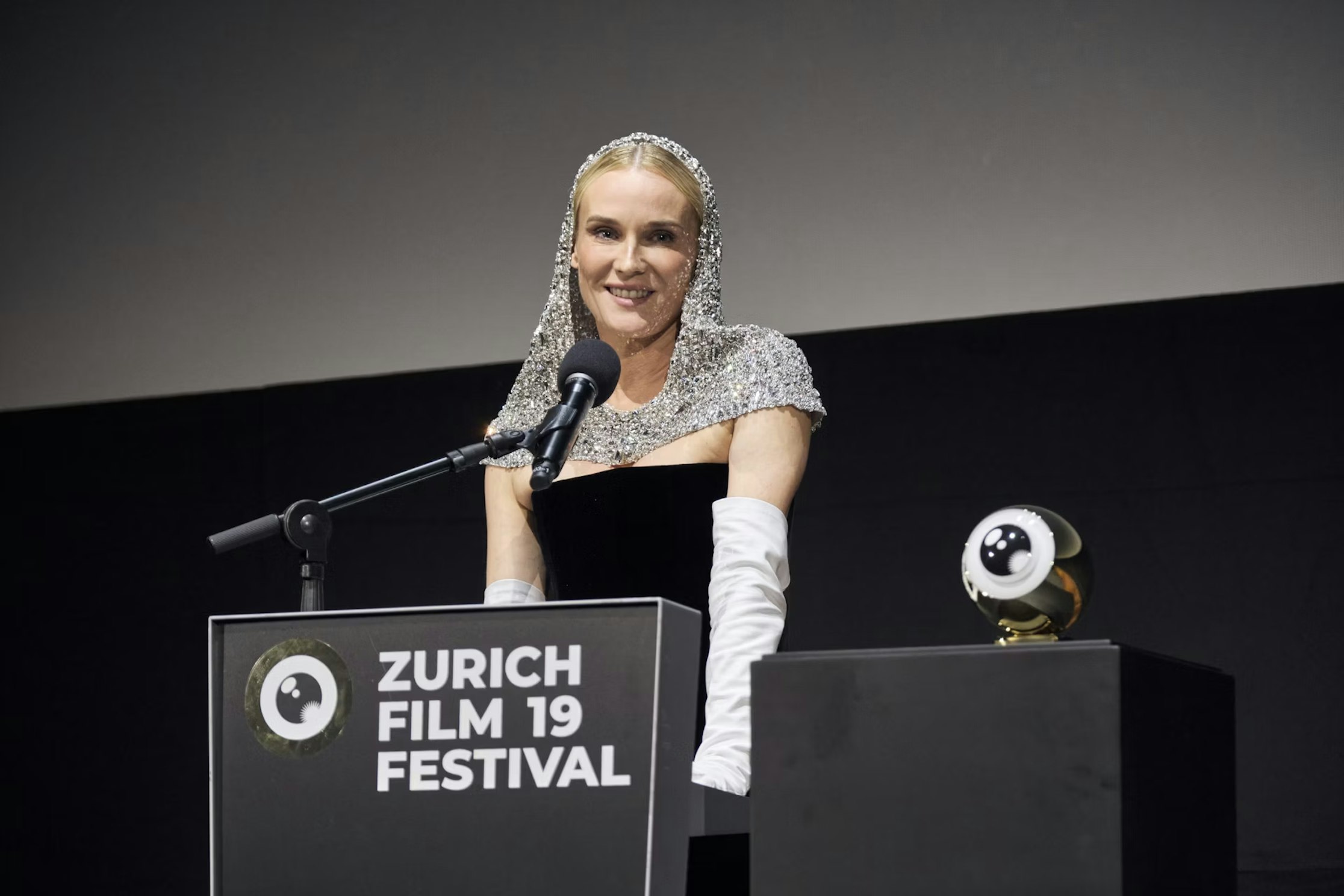 Eine Frau in einem glitzernden Kapuzenkostüm und weißen Handschuhen steht lächelnd mit einem Mikrofon an einem Podium beim Zurich Film Festival. Auf einem Podest in der Nähe steht eine Trophäe in Form eines Auges.