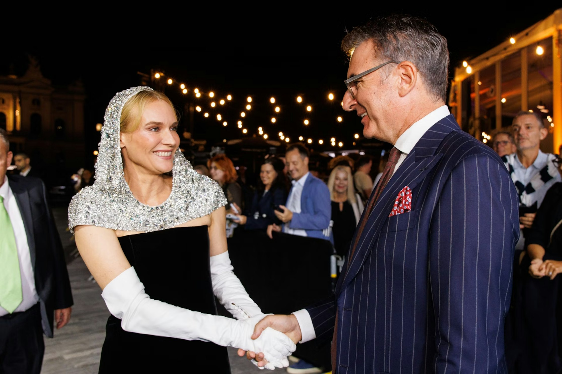 Eine Frau in einem glitzernden Kapuzenkleid und weißen Handschuhen schüttelt einem Mann im Nadelstreifenanzug bei einer nächtlichen Veranstaltung die Hand, im Hintergrund sind Lichterketten und eine Menschenmenge zu sehen.