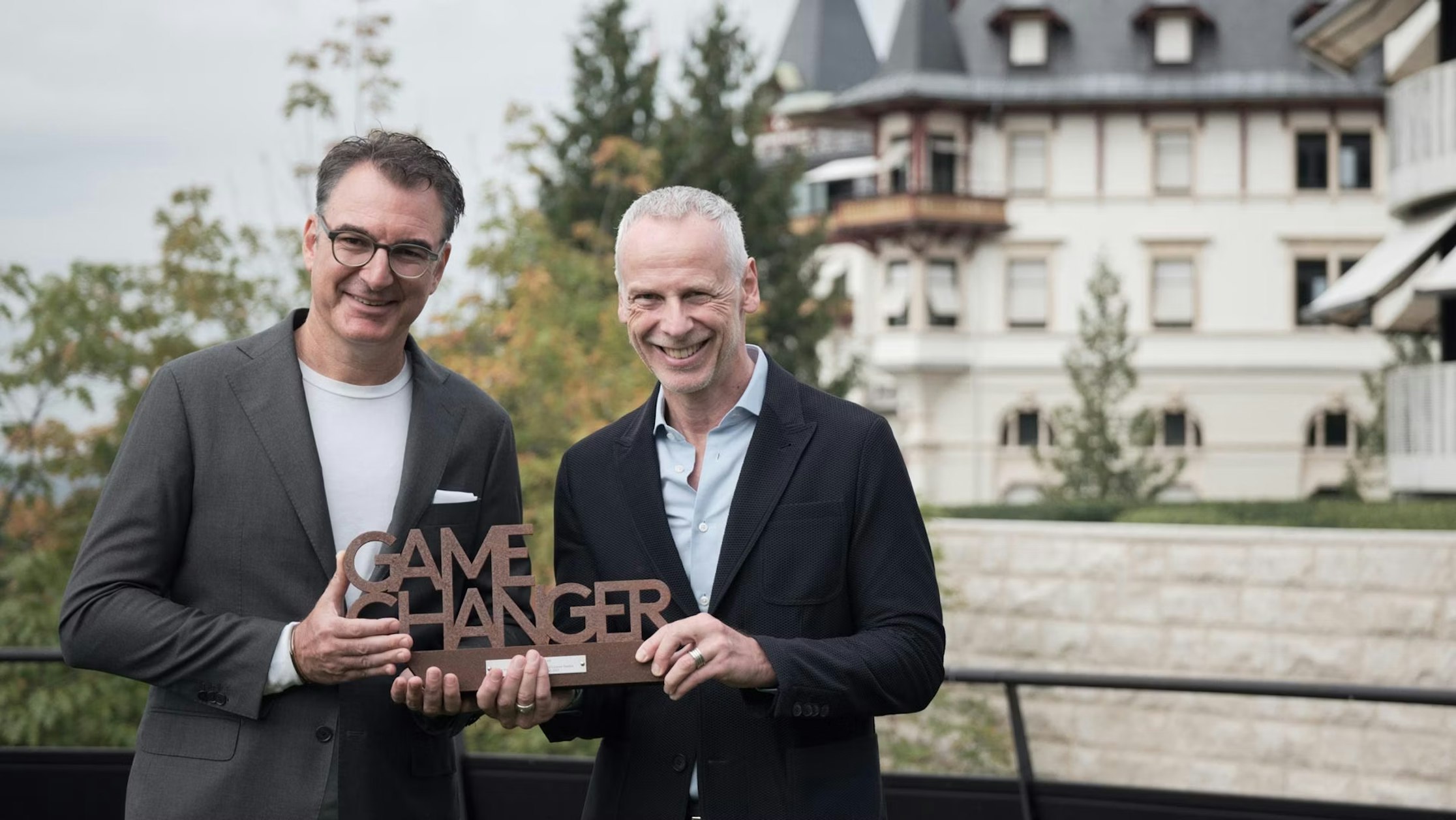 Zwei Männer in Anzügen stehen im Freien vor einem großen Gebäude, lächeln und halten gemeinsam ein hölzernes „GAME CHANGER“-Schild. Im Hintergrund sind Bäume und ein Teil des Gebäudes zu sehen.