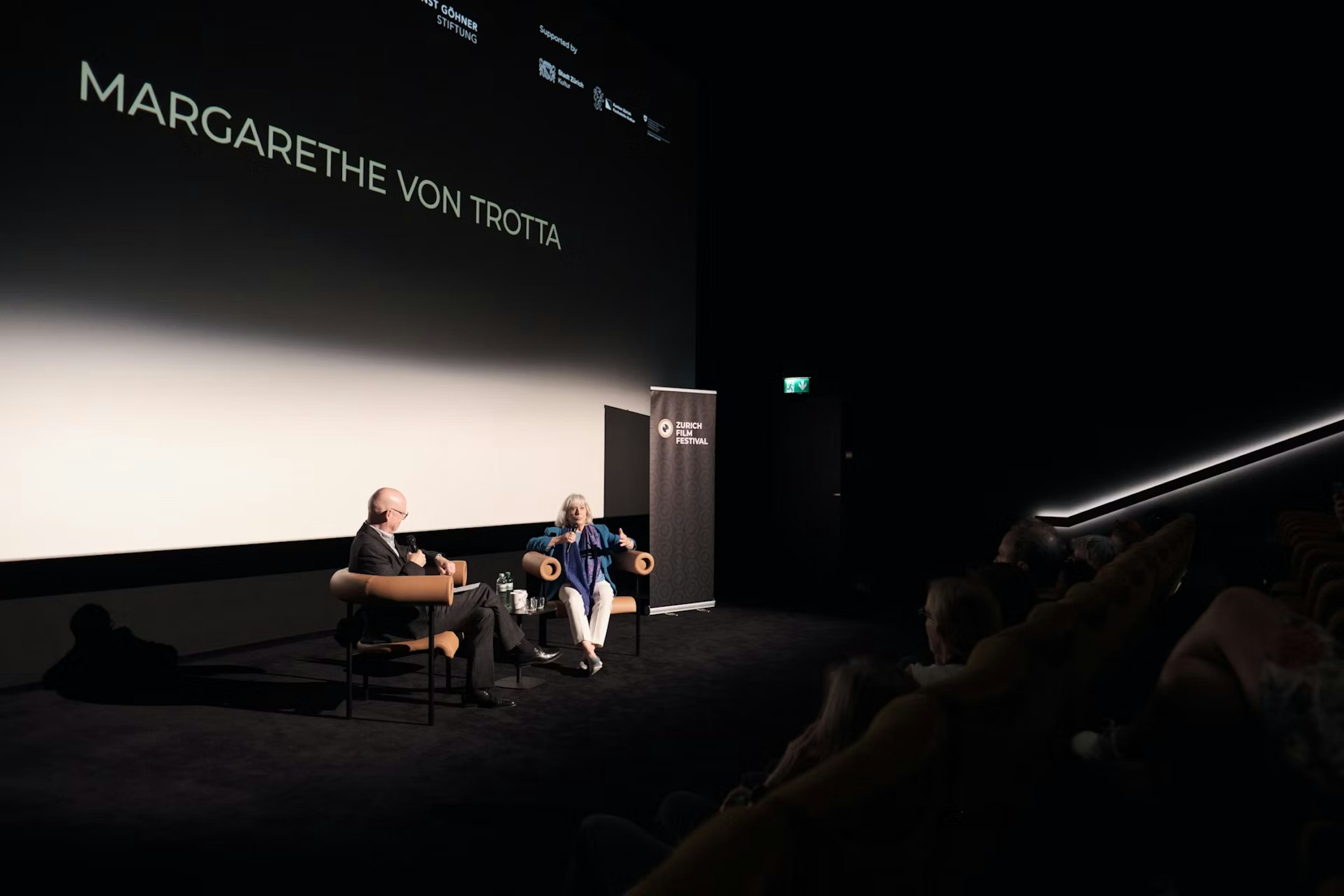 Zwei Personen sitzen in Sesseln auf der Bühne und unterhalten sich in einem dunklen Theater. Auf der Leinwand hinter ihnen wird der Name Margarethe von Trotta projiziert. Im Vordergrund sind Zuschauer zu sehen.