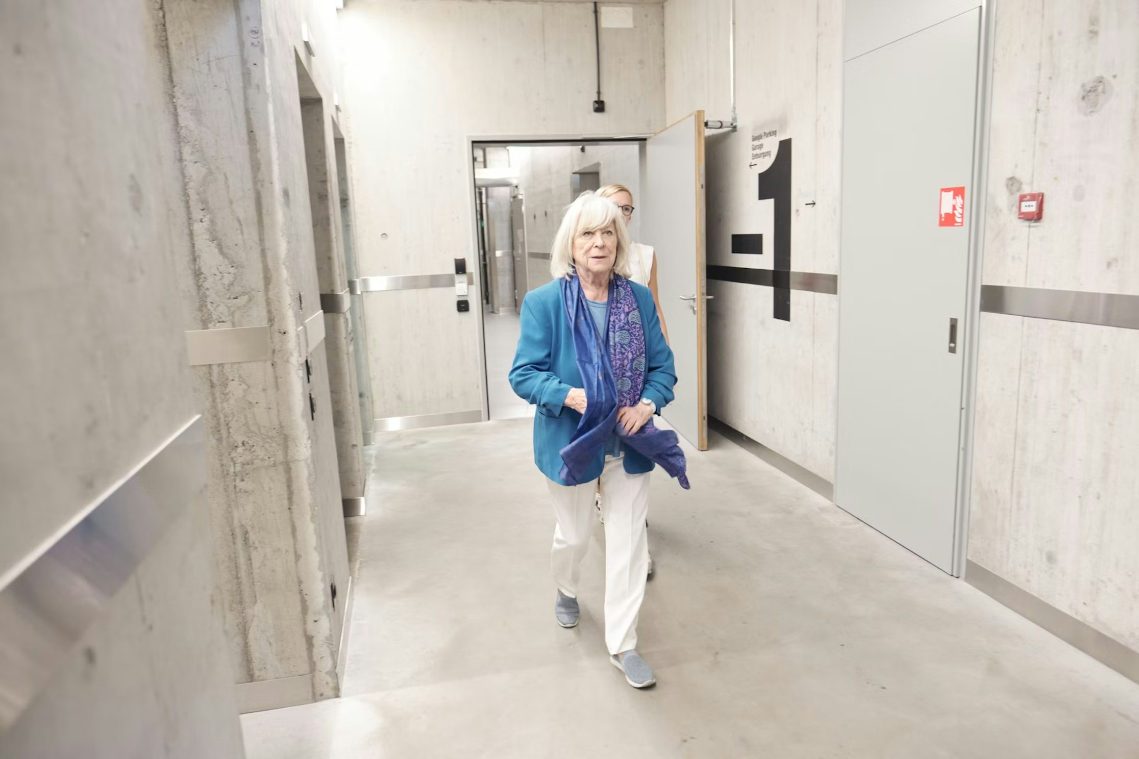 Eine ältere Frau mit weißem Haar, die eine blaue Jacke und weiße Hosen trägt, geht einen Betonflur entlang, eine andere Person folgt ihr.