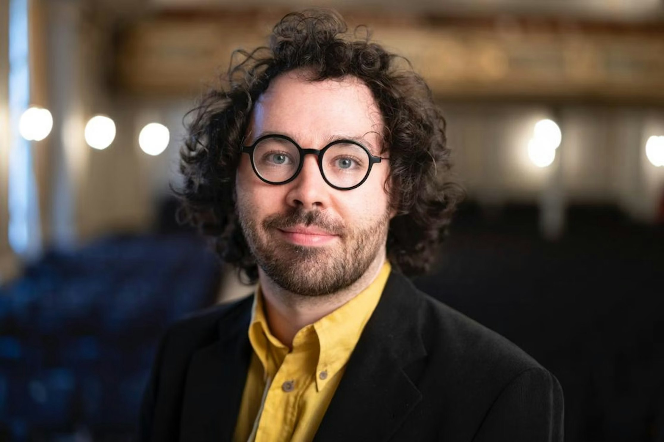 Ein Mann mit lockigem braunem Haar, Bart und runder Brille, der einen schwarzen Blazer über einem gelben Hemd trägt, steht in einem Gebäude mit verschwommenem Licht und Stühlen im Hintergrund.