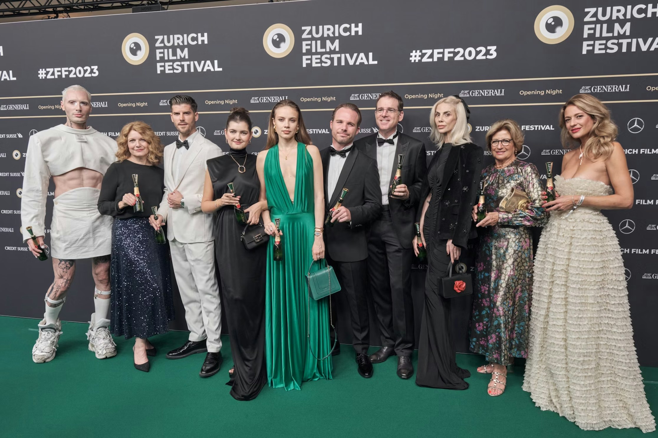 Eine Gruppe von zehn Personen in formellen und kreativen Outfits posiert gemeinsam auf dem grünen Teppich des Zurich Film Festival 2023, jeder hält eine Trophäe in den Händen. Im Hintergrund sind Festivallogos und Sponsoren zu sehen.