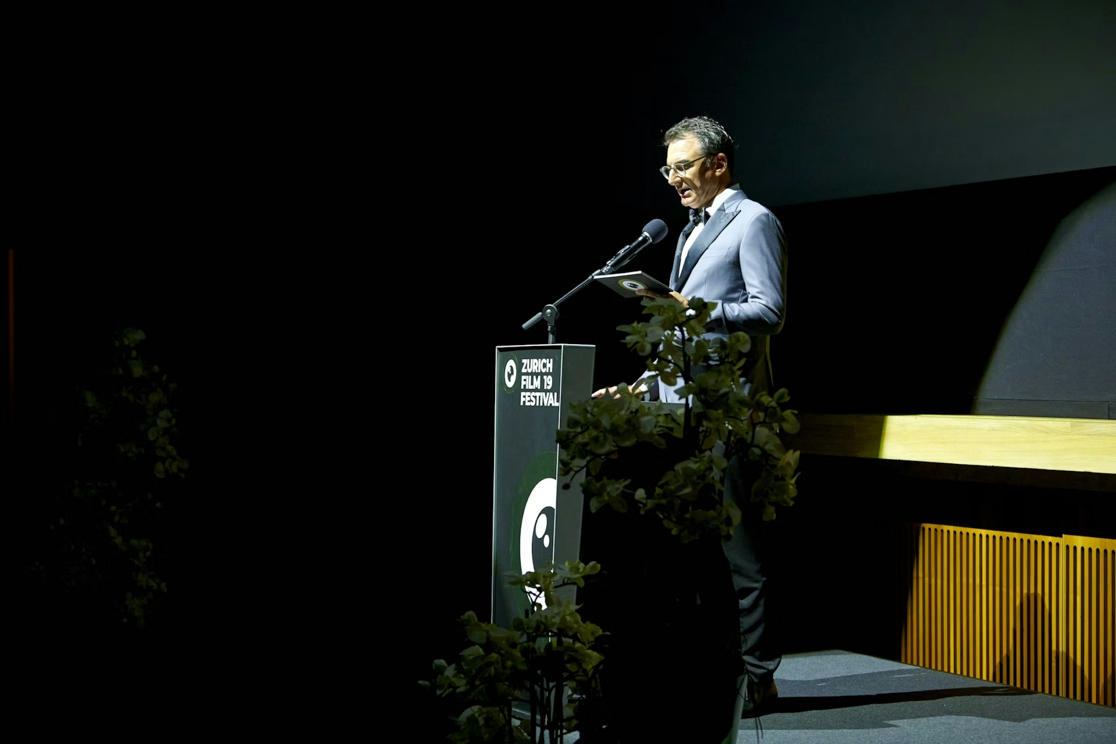 Ein Mann im Anzug steht auf einem Podium und spricht in ein Mikrofon auf der schwach beleuchteten Bühne des Zurich Film Festival. Auf dem Podium sind Blumen und das Festivallogo zu sehen.