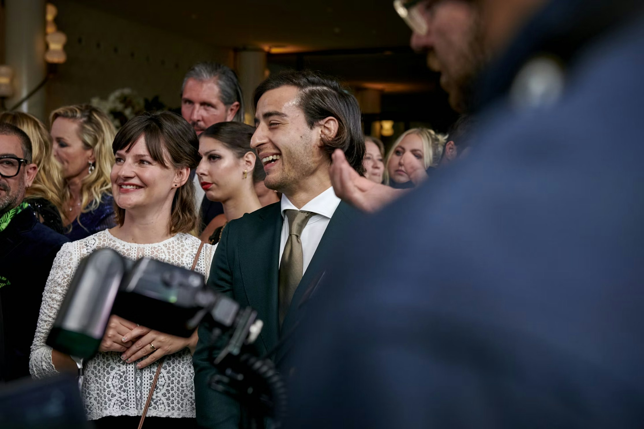 Eine Gruppe gut gekleideter Menschen, darunter ein lächelnder Mann im Anzug und eine Frau in einem weißen Spitzentop, stehen bei einer Veranstaltung in geschlossenen Räumen zusammen, während andere im Hintergrund zuschauen und sich unterhalten.