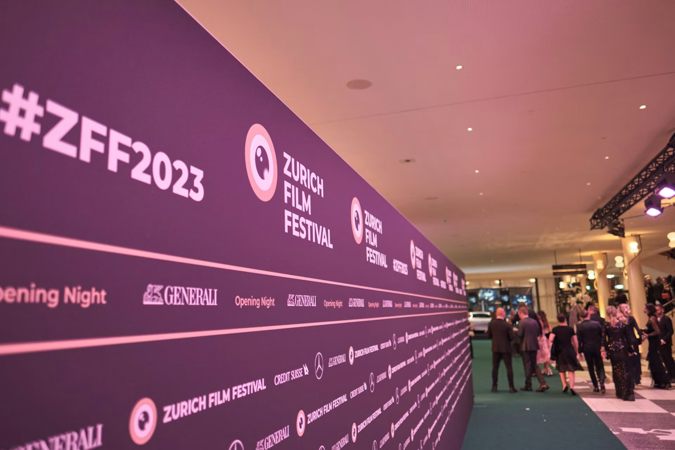 Auf einem violetten Hintergrund mit sich wiederholenden Motiven sind „Zurich Film Festival“ und „#ZFF2023“ zu sehen. Menschen in formeller Kleidung versammeln sich auf einem grünen Teppich in einem hell erleuchteten Veranstaltungsort.