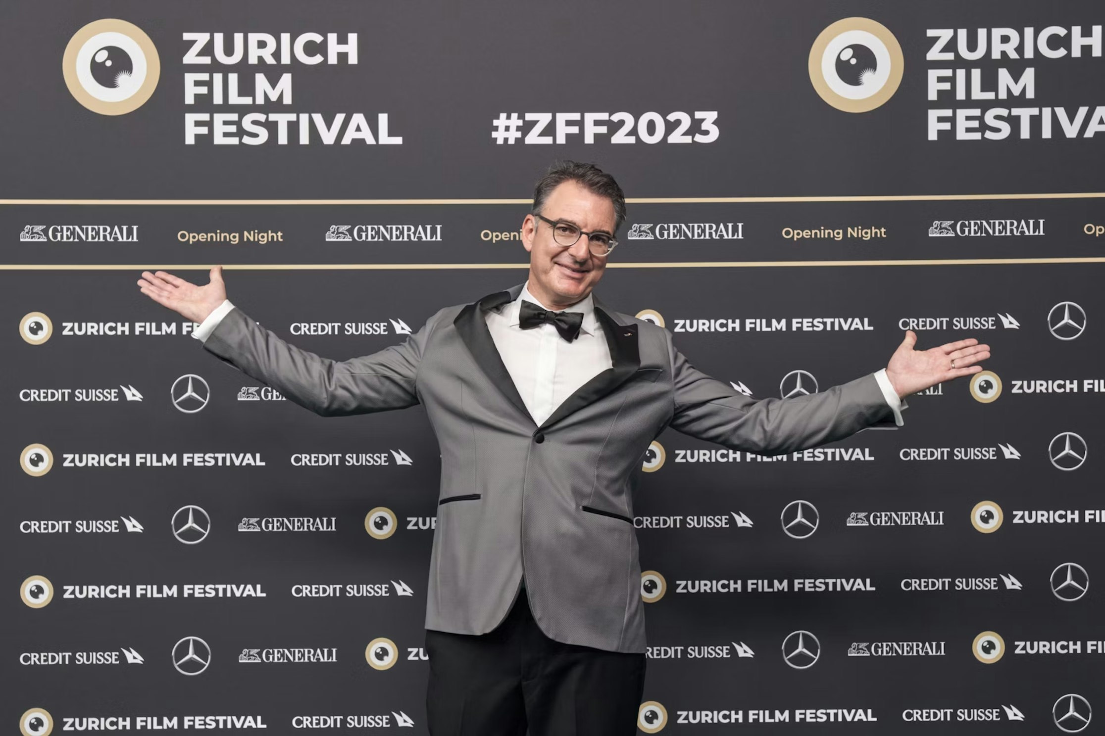 Ein Mann in silbernem Smoking und Fliege steht lächelnd und mit weit ausgebreiteten Armen auf dem roten Teppich des Zurich Film Festival, vor einem Hintergrund mit Sponsorenlogos und #ZFF2023.