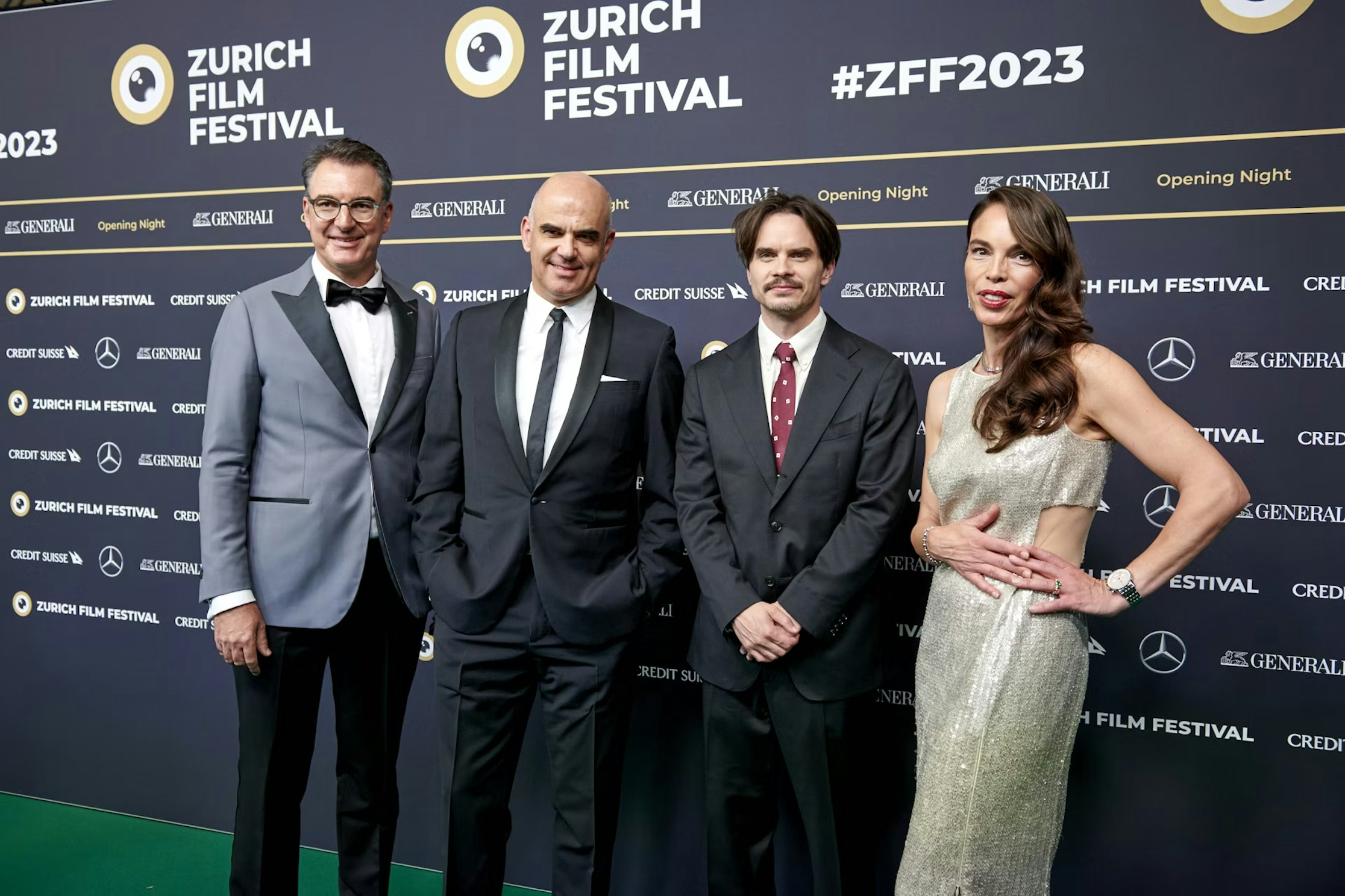 Vier Personen in formeller Kleidung stehen auf einem grünen Teppich und posieren für ein Foto beim Zurich Film Festival, im Hintergrund sind Event-Banner mit Markenzeichen zu sehen.