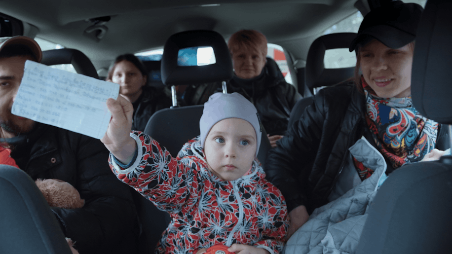 Ein kleines Kind in einer bunten Jacke hält eine handgeschriebene Notiz hoch, während es auf dem Rücksitz eines Autos sitzt, umgeben von vier Erwachsenen, die mit neutralem oder freundlichem Gesichtsausdruck nach vorne blicken.