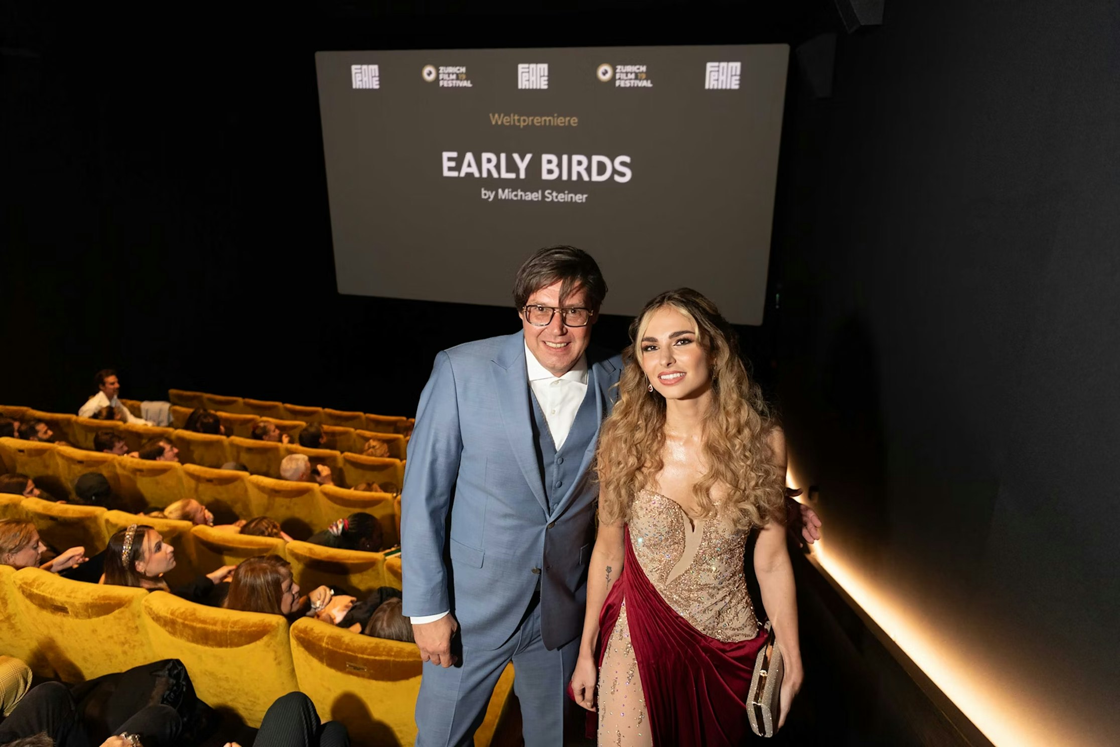 Ein Mann im blauen Anzug und eine Frau im roten Abendkleid posieren und lächeln vor einem Kinopublikum, während auf der Leinwand bei einer Filmpremiere „EARLY BIRDS“ von Michael Steiner läuft.