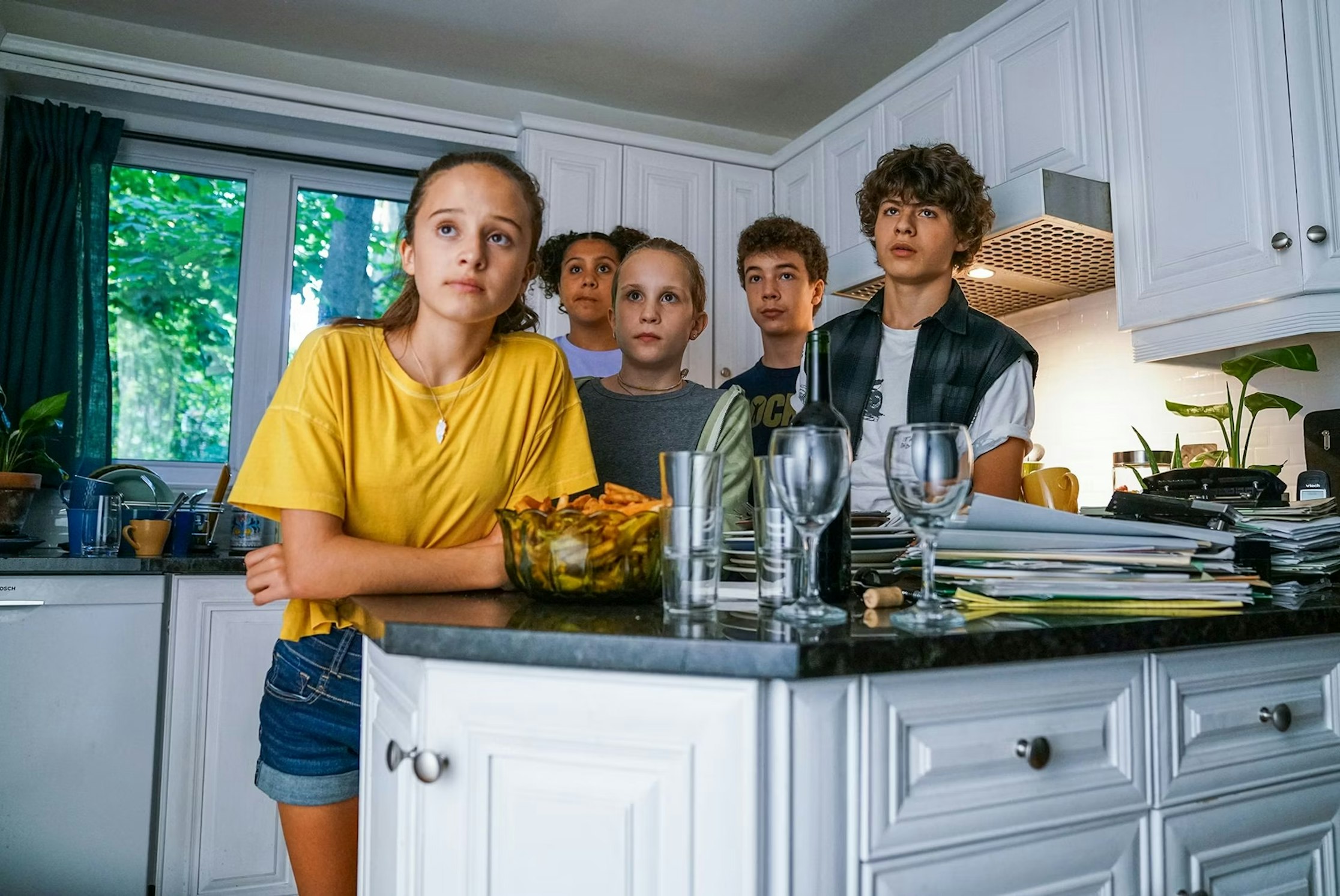 Fünf Kinder stehen eng beieinander in einer Küche und schauen mit ernstem Blick geradeaus. Die Küche hat weiße Schränke, ein Fenster mit Blick ins Grüne und eine Arbeitsplatte mit Gläsern und einer Schüssel mit Snacks.