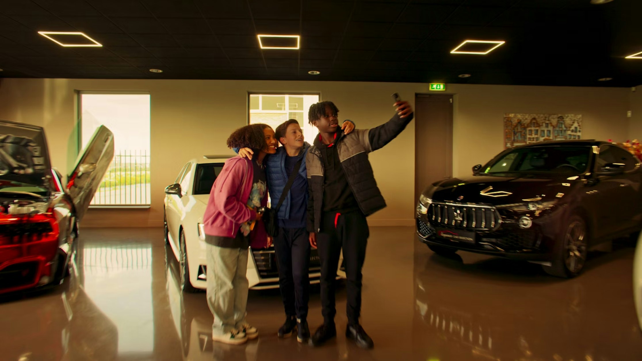 Drei Personen stehen dicht beieinander in einem Autohaus, lächeln und machen ein Selfie. Zu sehen sind zwei Luxusautos, eines weiß mit offener Tür und eines schwarz, beide auf glänzendem Boden.