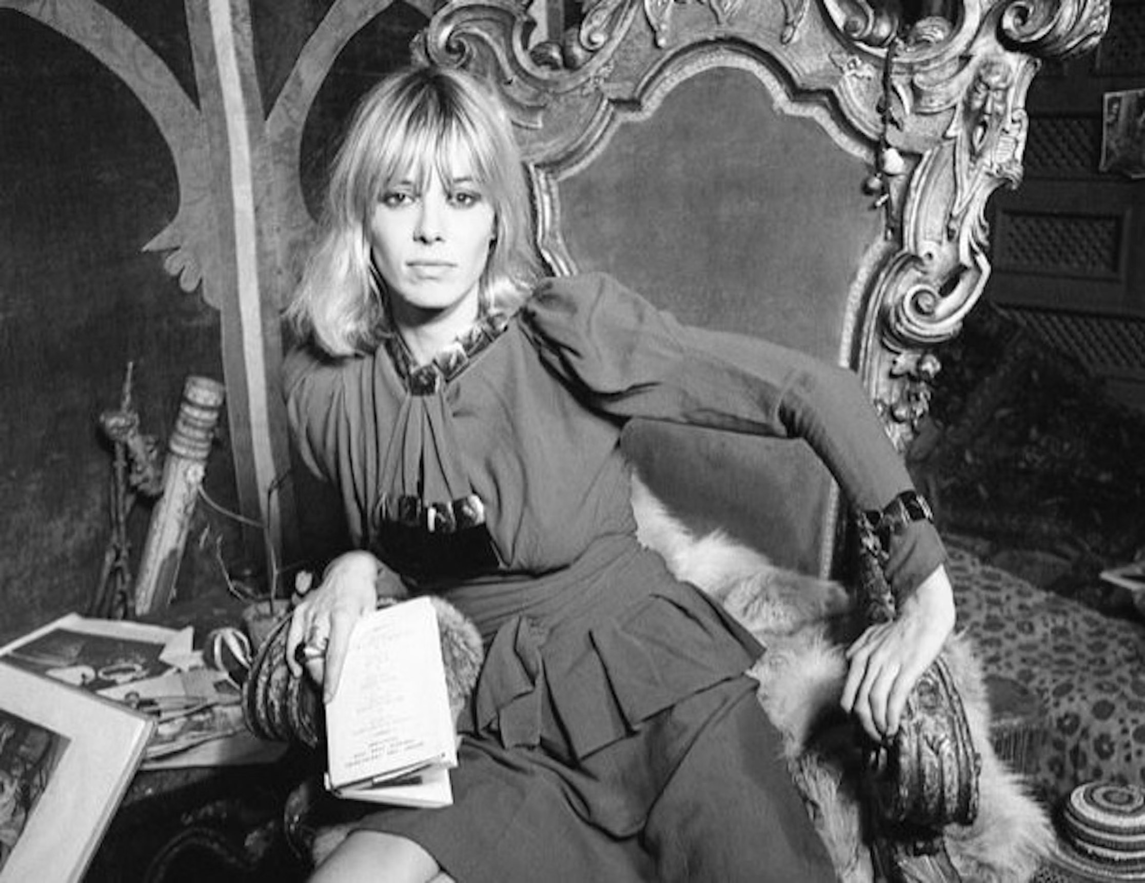 Eine Frau mit blondem Haar und einem Vintage-Kleid sitzt auf einem kunstvoll geschnitzten Stuhl und hält ein Buch oder eine Zeitung in der Hand, umgeben von eklektischem Dekor und gemusterten Möbeln.