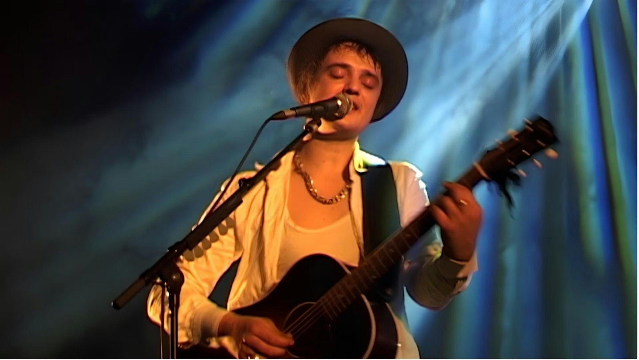 Eine Person mit Hut und weißem Hemd spielt auf der Bühne eine Akustikgitarre und singt in ein Mikrofon, im Hintergrund ist eine dramatische blaue und gelbe Beleuchtung zu sehen.