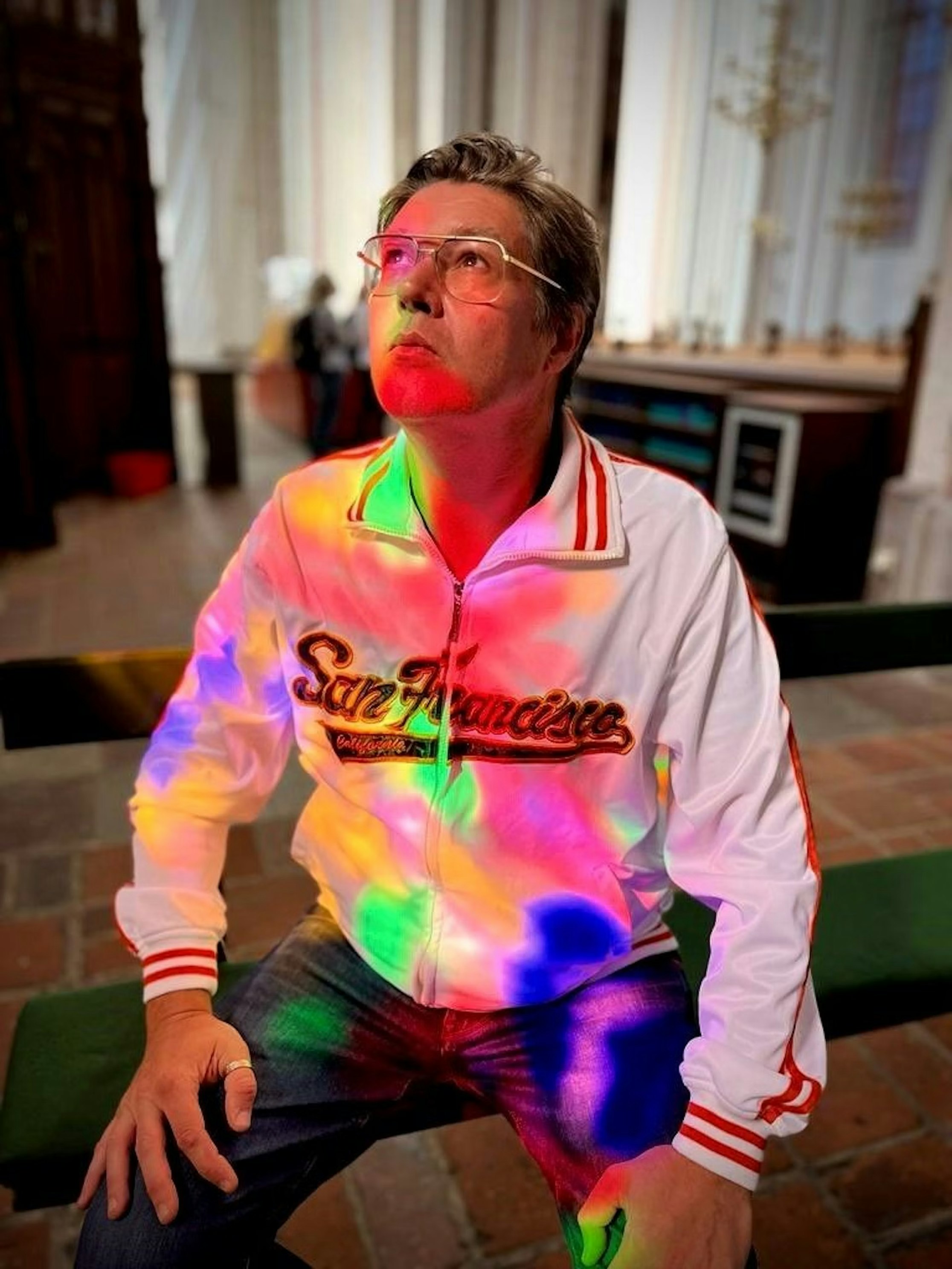 Ein Mann mit Brille und weißer San Francisco-Jacke sitzt im Haus und blickt nach oben, während bunte Lichter in Rot, Grün, Blau und Lila auf sein Gesicht und seine Kleidung leuchten. Der Hintergrund ist von Holz und Stein geprägt.