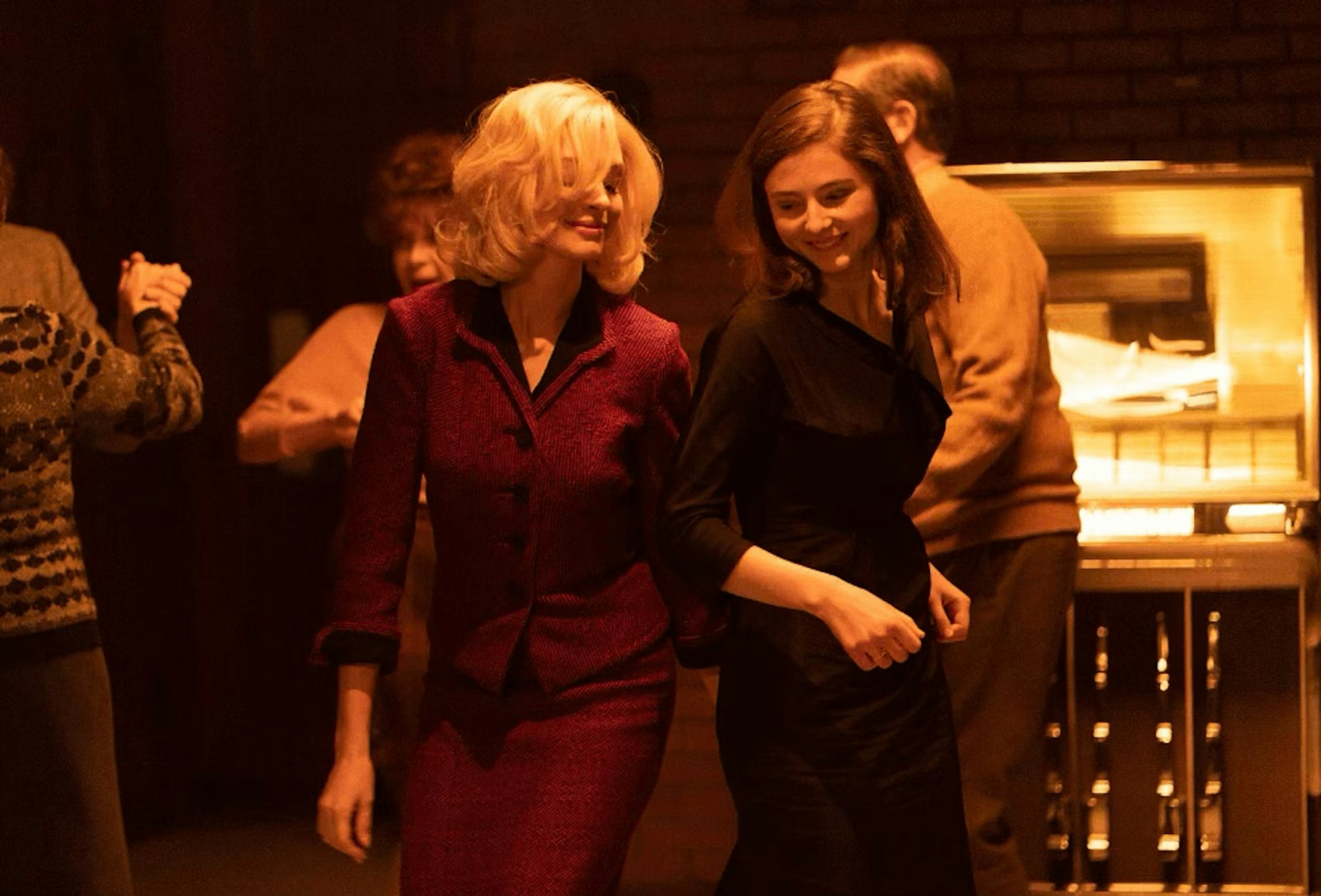 Zwei Frauen, eine im roten Anzug und die andere im schwarzen Kleid, lächeln und gehen zusammen in einem warm beleuchteten Raum mit einer Jukebox und anderen tanzenden Menschen im Hintergrund.
