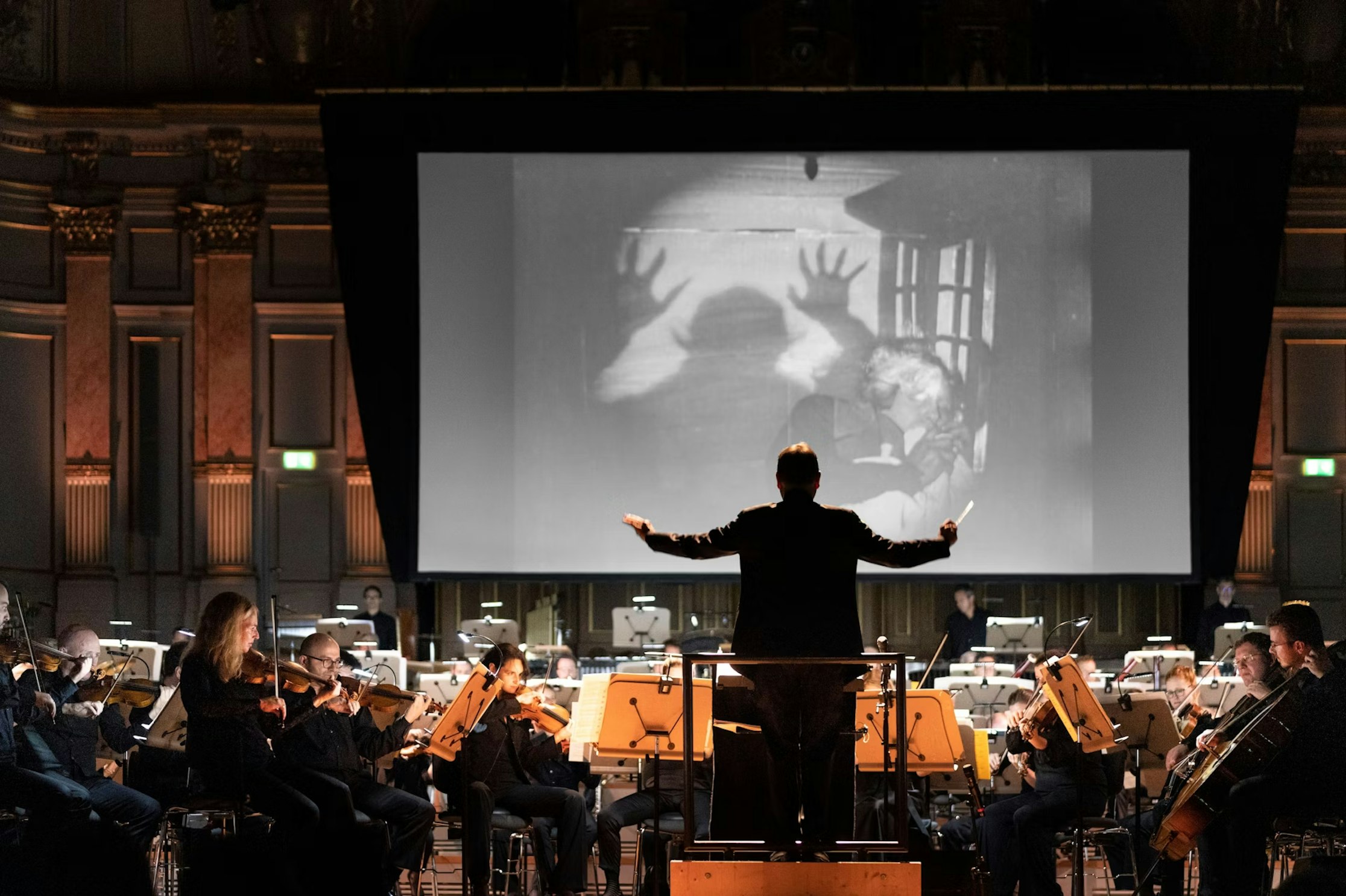 Ein Dirigent leitet ein Orchester, das vor einer großen Leinwand spielt. Auf der Leinwand ist eine Schwarz-Weiß-Szene aus einem Horrorfilm zu sehen, in der schattenhafte Hände nach einer verängstigten Person greifen. Die Musiker sitzen auf der Bühne eines großen Konzertsaals.