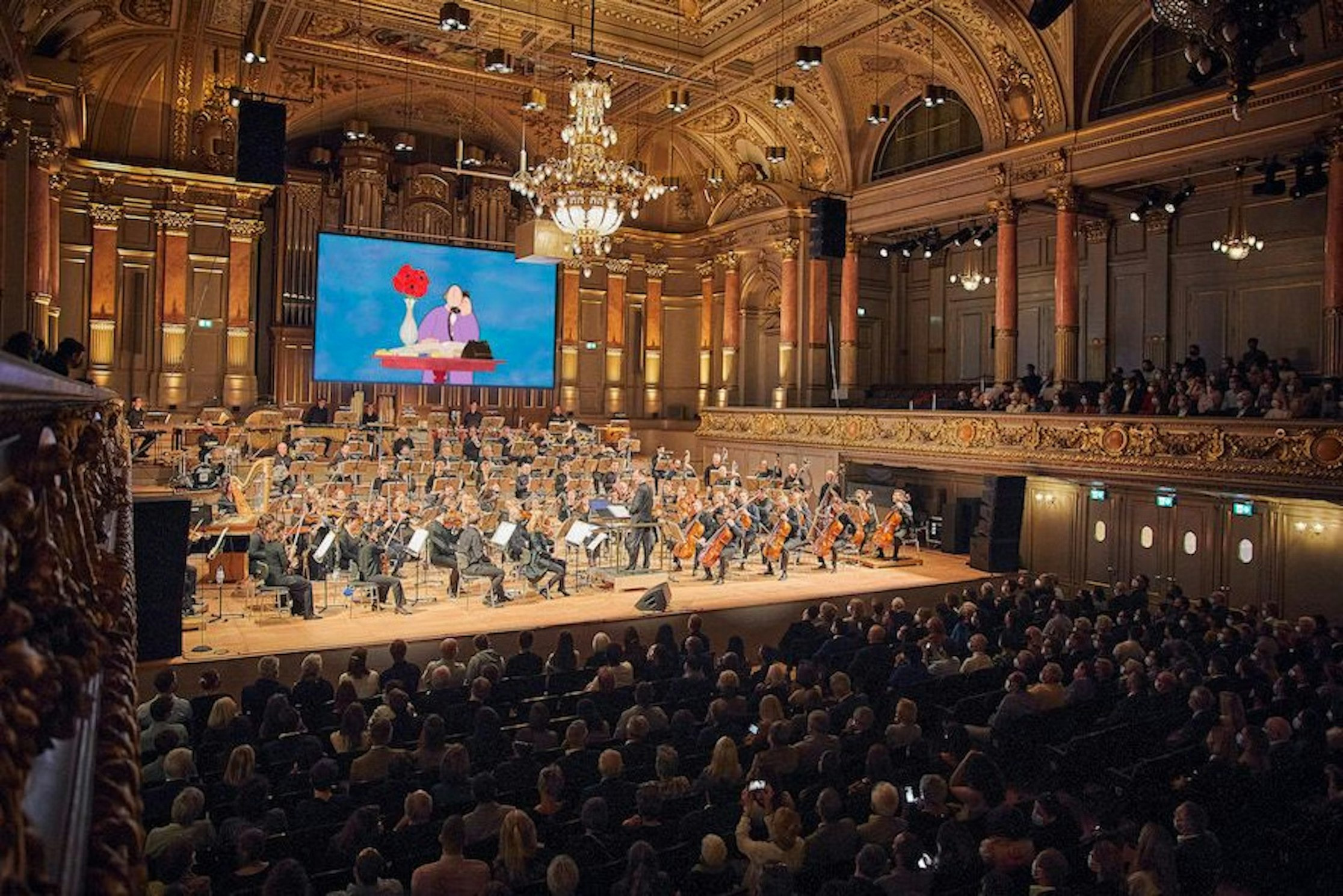 Ein großes Orchester spielt in einem großen, mit Publikum gefüllten Konzertsaal. Über den Musikern zeigt ein Bildschirm eine animierte Szene mit zwei Figuren und einer roten Blume. Der Saal ist mit Kronleuchtern und Balkonen geschmückt.