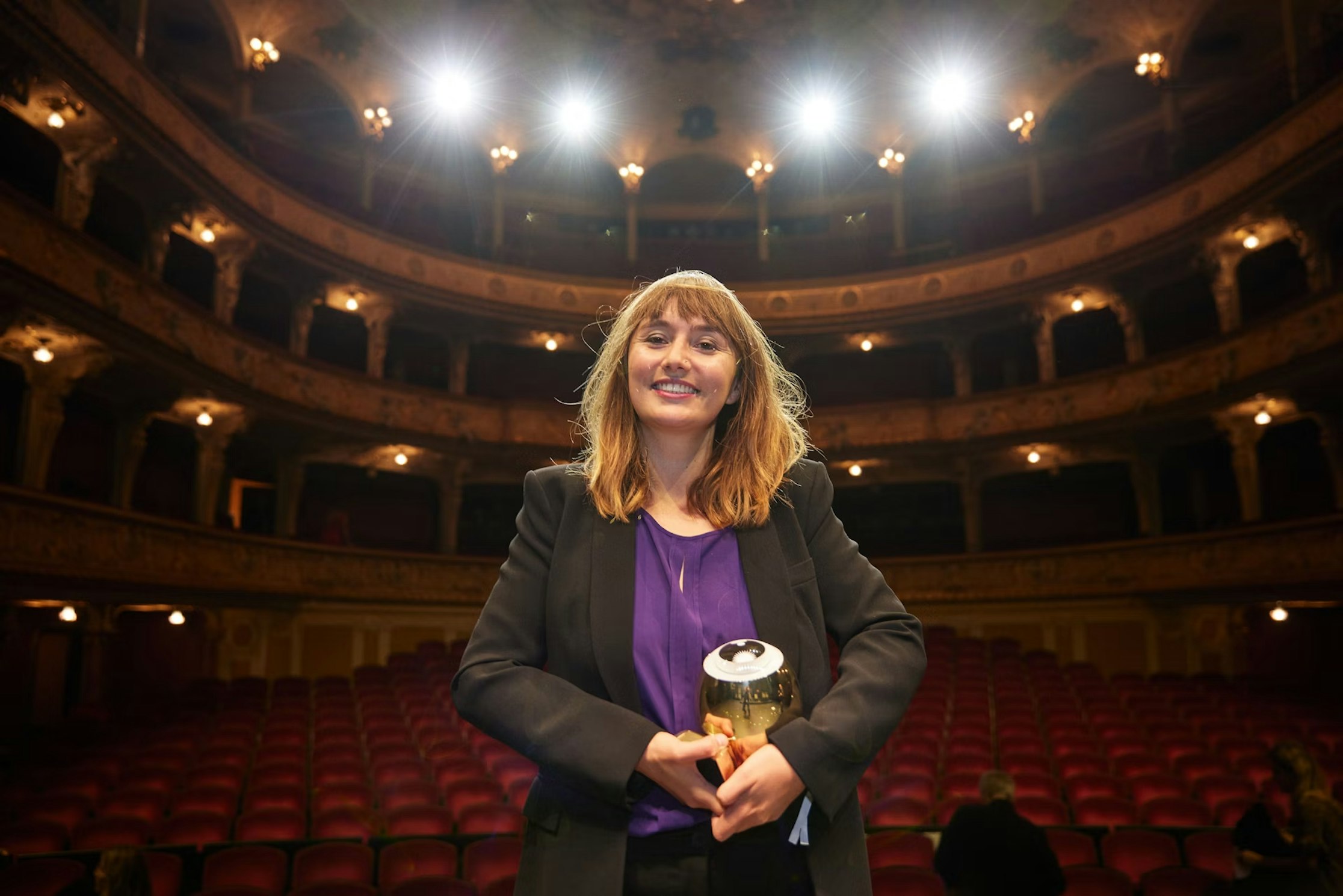 Eine Frau in einem schwarzen Blazer und einem lila Hemd steht lächelnd und mit einer Trophäe in der Hand auf der Bühne eines großen Theaters, hinter ihr leuchten helle Lichter und im Hintergrund sind leere rote Sitze zu sehen.