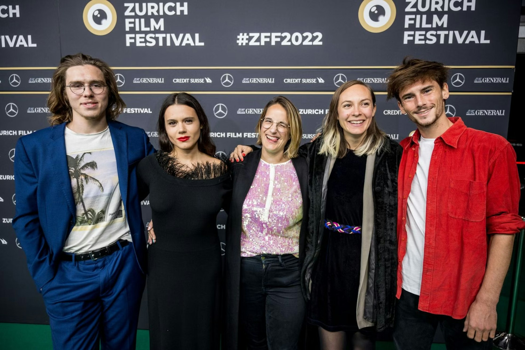 Fünf Personen stehen lächelnd auf dem grünen Teppich des Zurich Film Festival 2022 und posieren vor einer schwarzen Kulisse mit Festival- und Sponsorenlogos. Sie wirken glücklich und tragen halbformelle Kleidung.