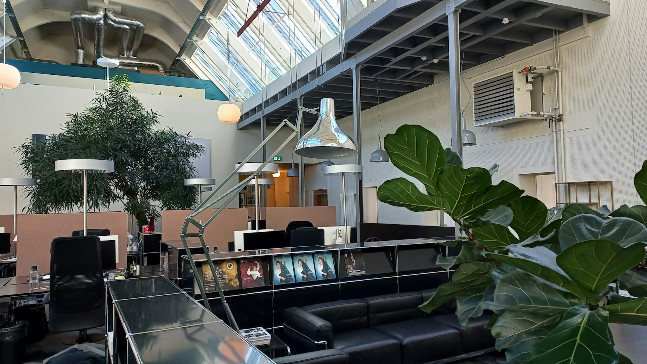 Moderne Büroräume mit hohen Decken, Oberlichtern und Beleuchtung im Industriestil. Es gibt Schreibtische mit Computern, schwarze Sofas, große Topfpflanzen und Poster an einer Trennwand. Die Atmosphäre ist offen und luftig.