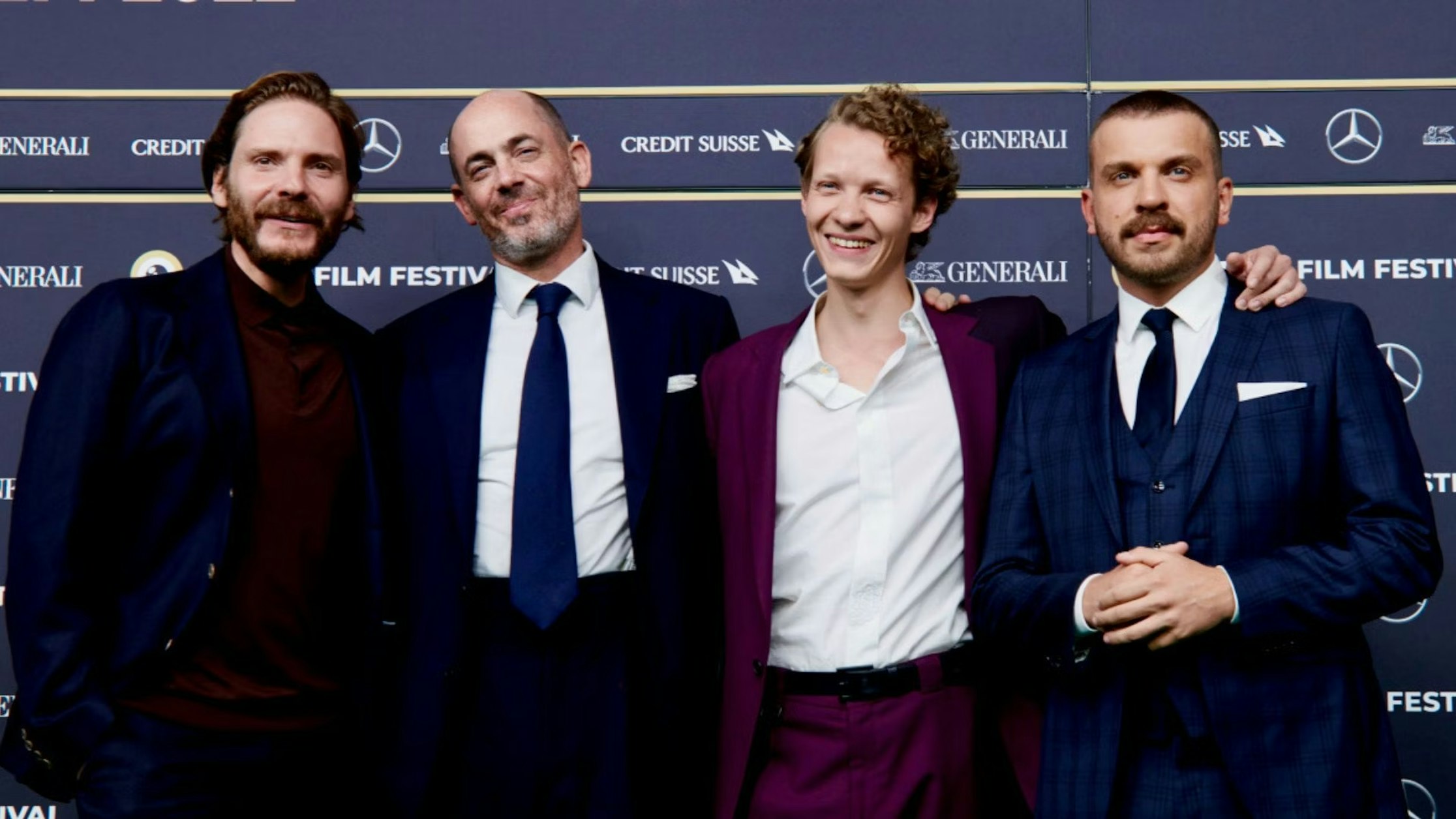 Vier Männer in Anzügen stehen zusammen, lächeln und posieren für ein Foto auf dem roten Teppich eines Filmfestivals, hinter ihnen sind die Logos der Markensponsoren zu sehen.