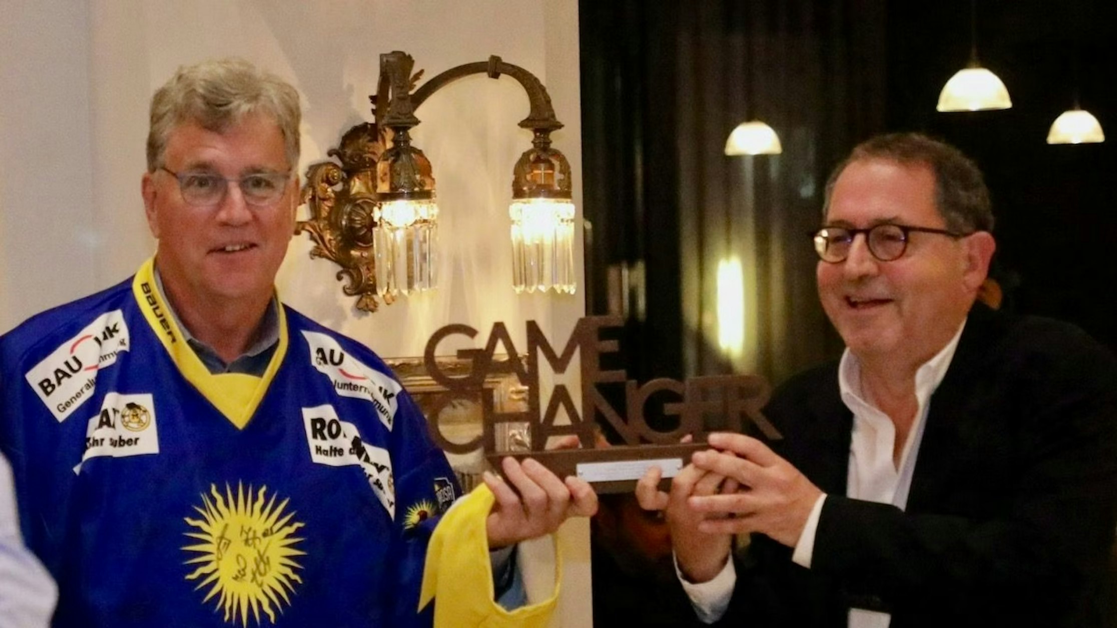 Zwei lächelnde Männer im Innenbereich; einer trägt ein blau-gelbes Sporttrikot, der andere einen dunklen Anzug. Beide halten einen Game Changer Award in den Händen. An der Wand hinter ihnen hängt eine Vintage-Lampe.