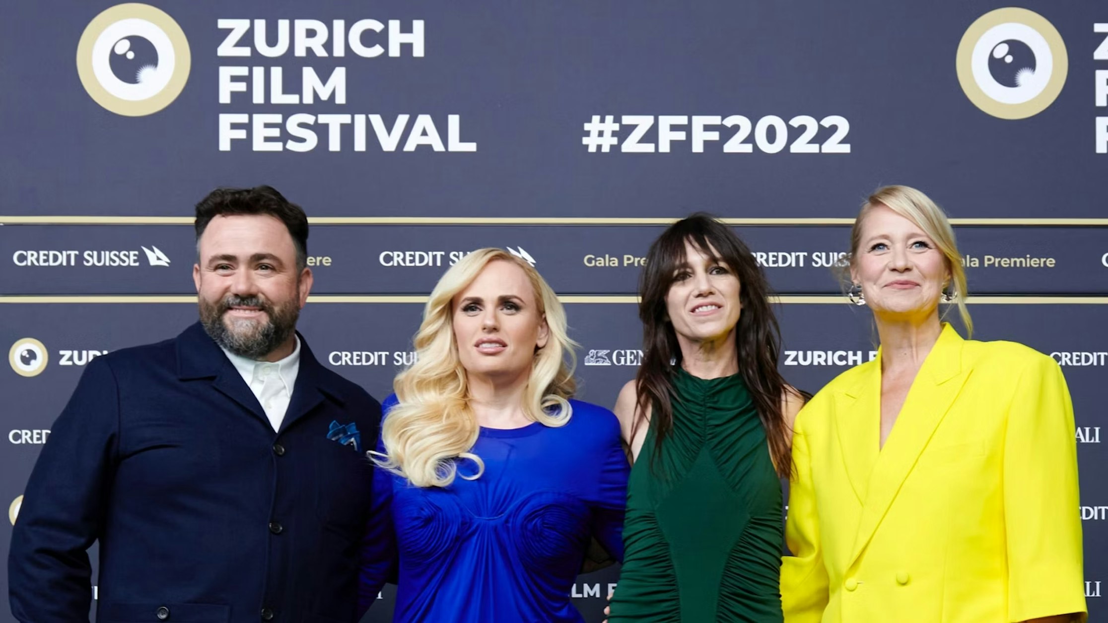 Vier Personen posieren gemeinsam vor der Kulisse des Zurich Film Festival. Alle tragen formelle Kleidung, darunter einen blauen Anzug, ein leuchtend blaues Kleid, ein grünes Kleid und einen leuchtend gelben Anzug.