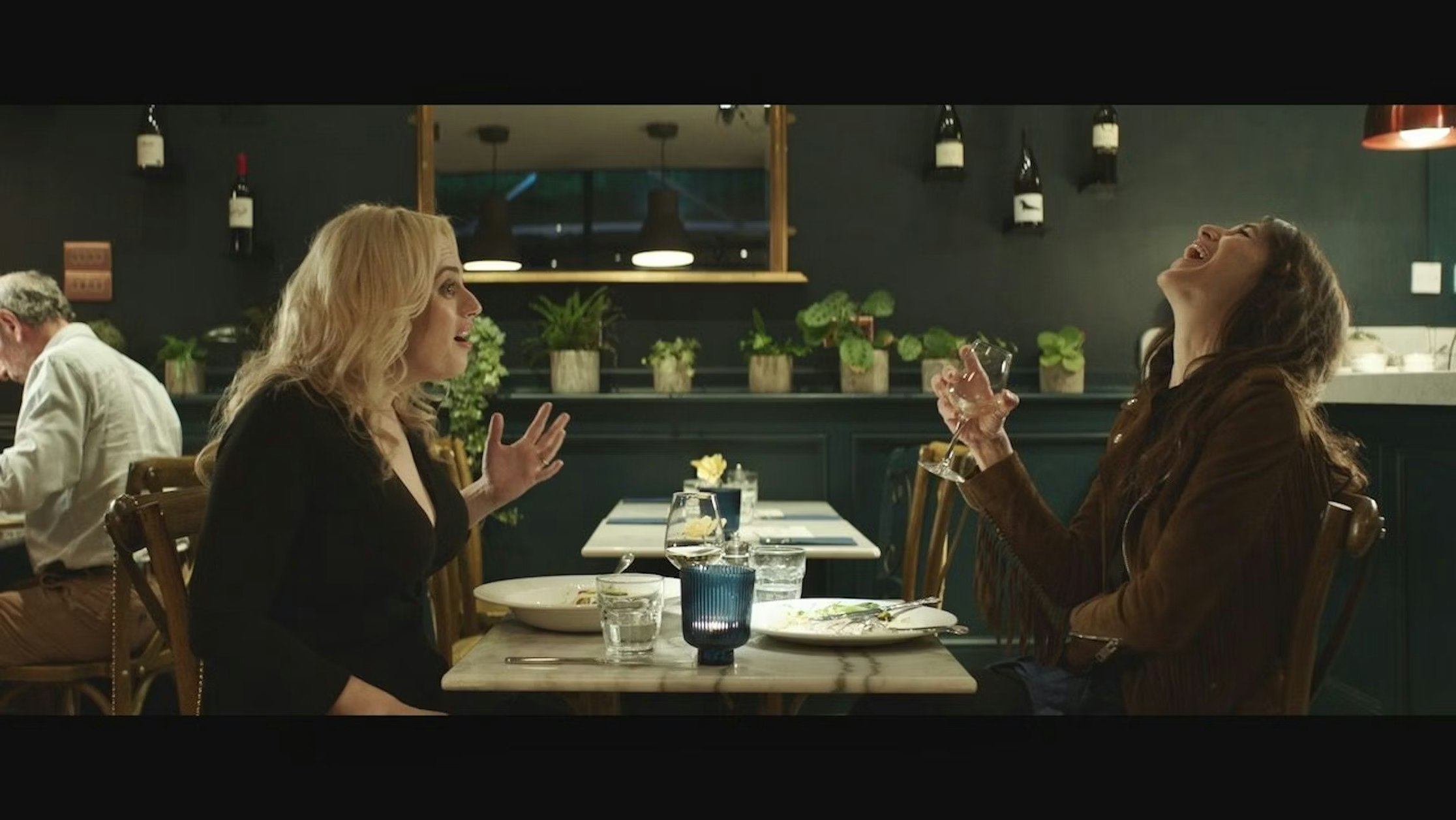 Zwei Frauen sitzen lachend und angeregt an einem Restauranttisch. Eine hält ein Glas Wein in der Hand. Auf dem Tisch stehen Teller, Gläser und eine Kerze, dahinter stehen Flaschen und Topfpflanzen auf Regalen.