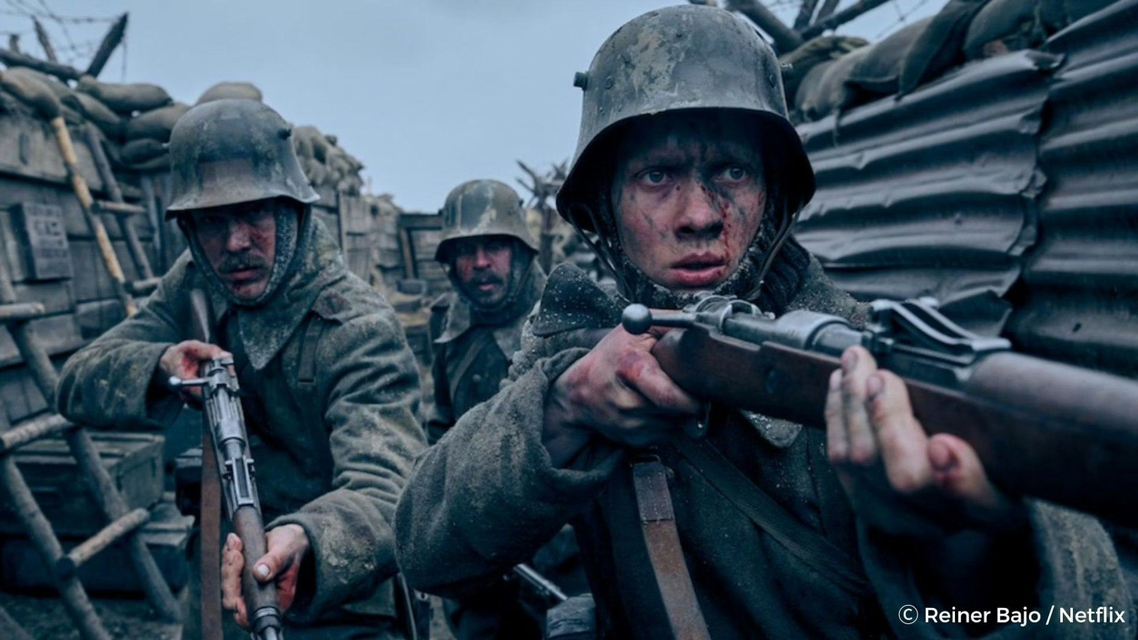 Drei Soldaten in schlammigen Uniformen und Helmen zielen mit Gewehren, während sie in einem Schützengraben aus Kriegszeiten kauern. Ihre Gesichter sind angespannt und schmutzig, umgeben von Sandsäcken und Stacheldraht unter einem düsteren Himmel.